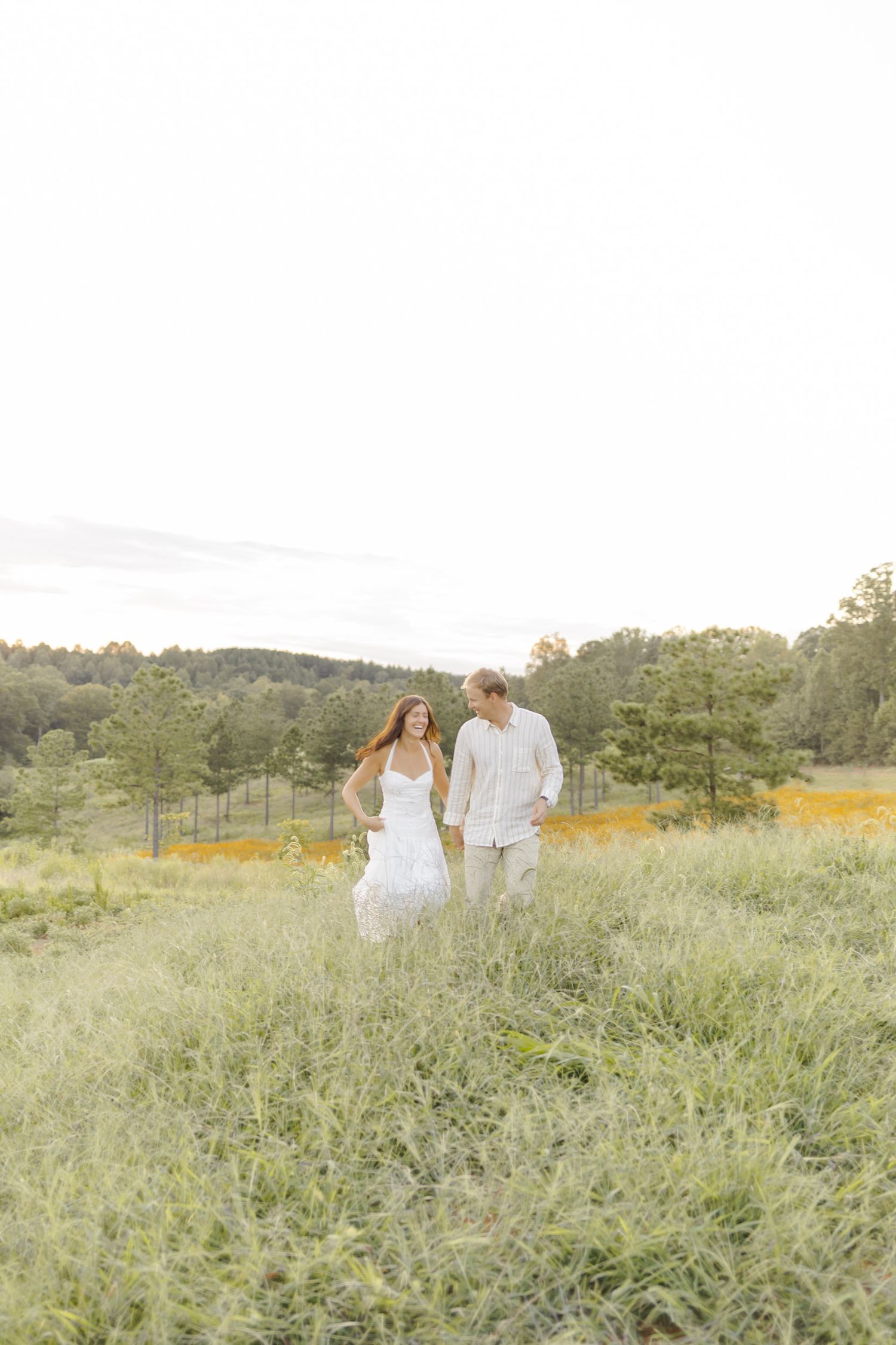 A couple in white attire walks hand-in-hand through a lush green meadow with trees in the background.
