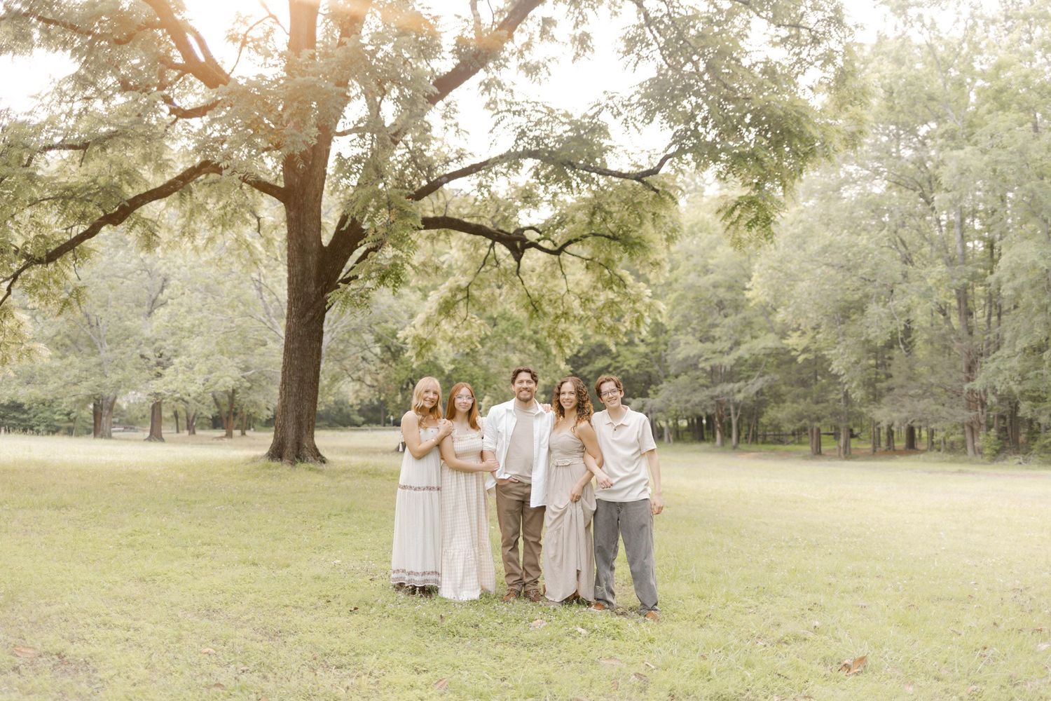 A group poses together in coordinated neutral outfits against a backdrop of trees in a park during lifestyle portrait photoshoot.
