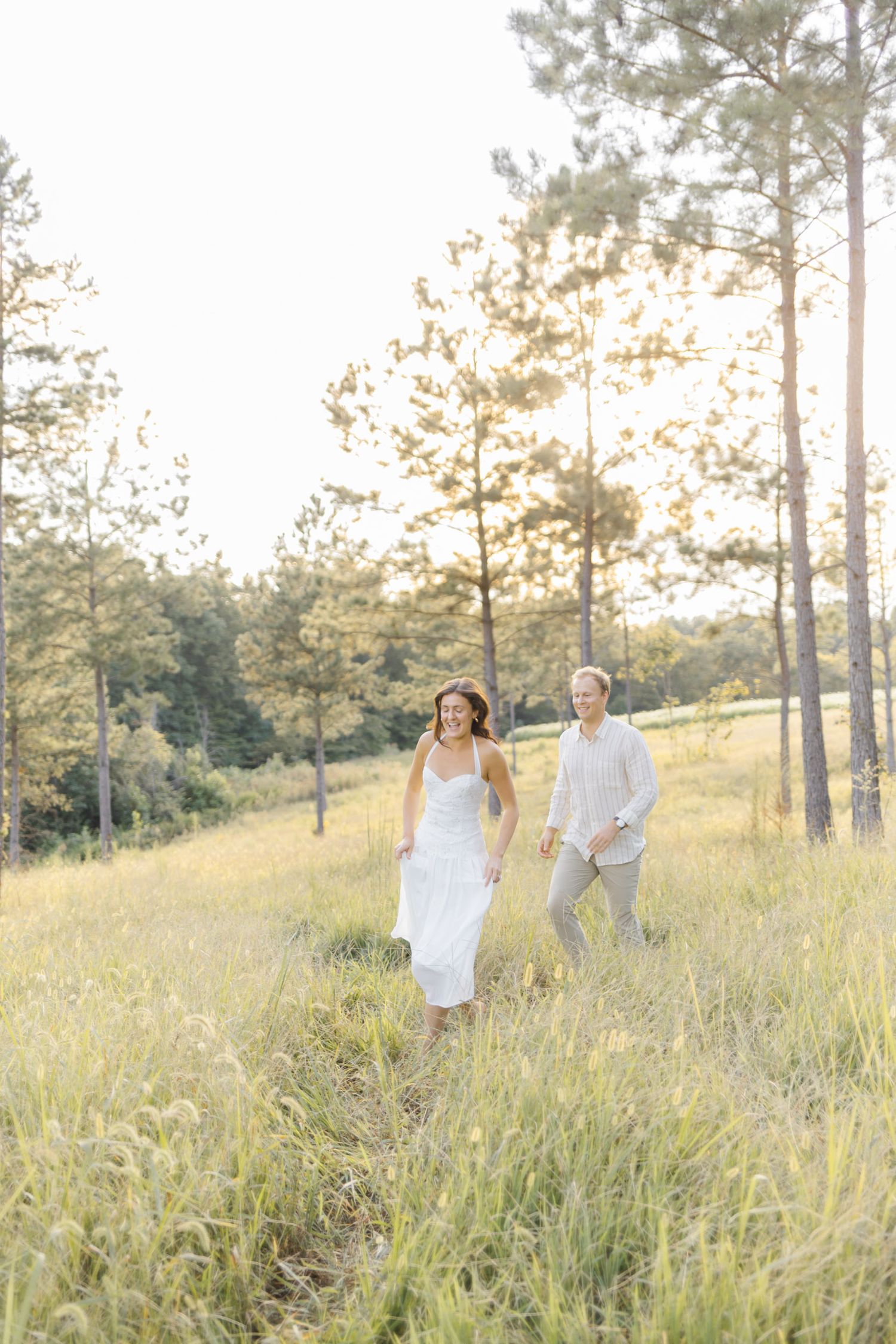 Couple dressed in white enjoying a peaceful moment in a grassy field surrounded by tall pines during lifestyle portrait photoshoot.