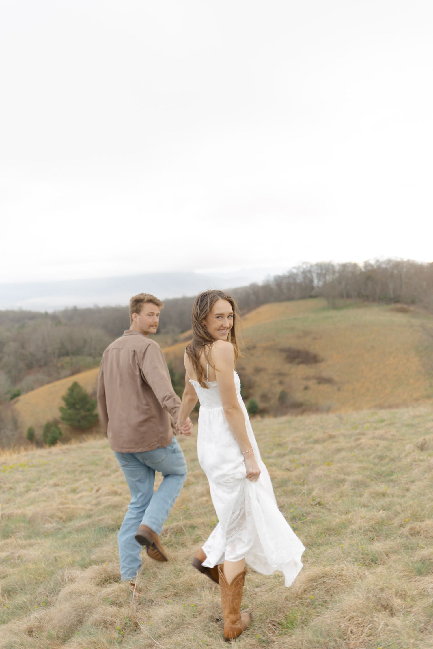 Couple walking hand-in-hand on a hilltop meadow, woman in flowing white dress and cowboy boots looks back at camera.