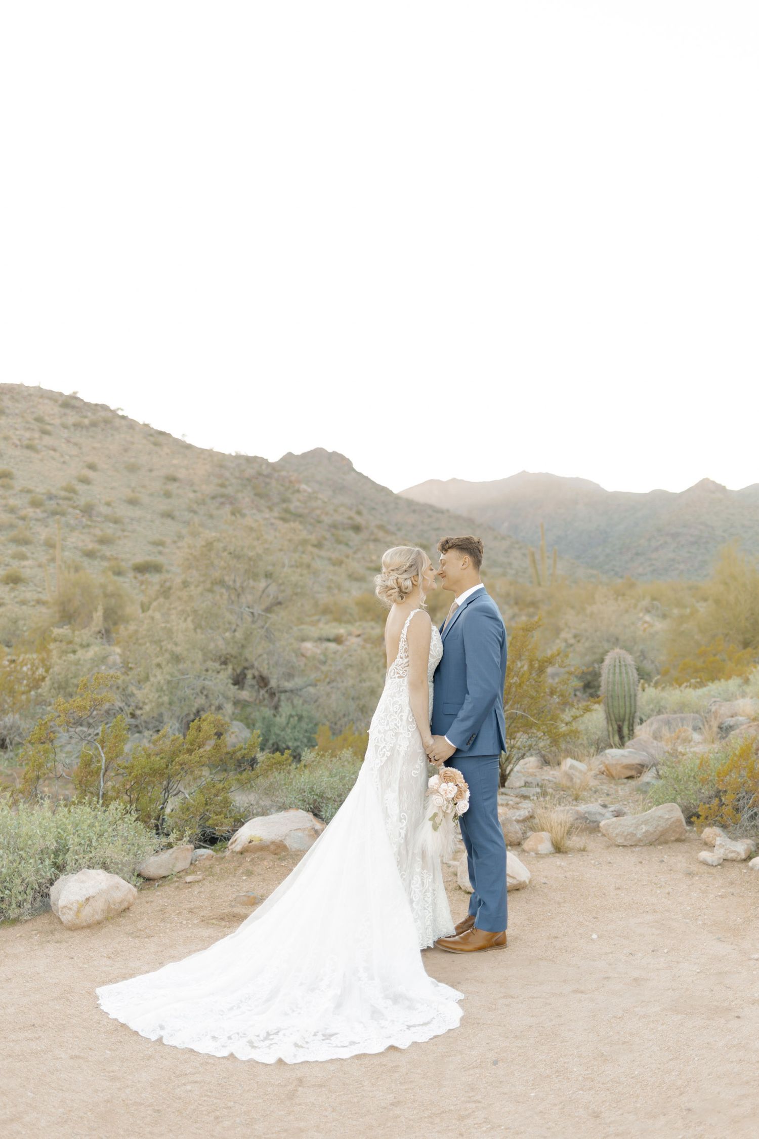 Bride and groom holding hands in desert landscape with saguaro cacti and mountains, bride wearing elegant lace gown with long train.