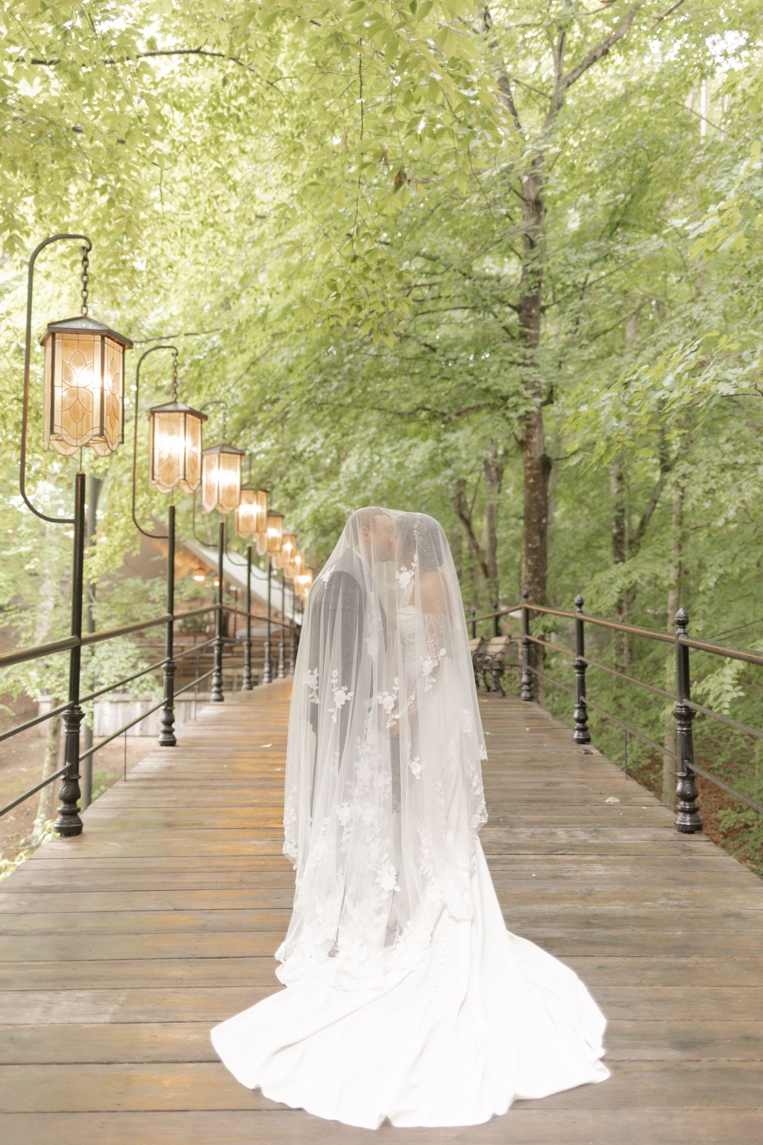 Bride in white gown and flowing veil stands on a lantern-lit wooden bridge surrounded by lush green trees.