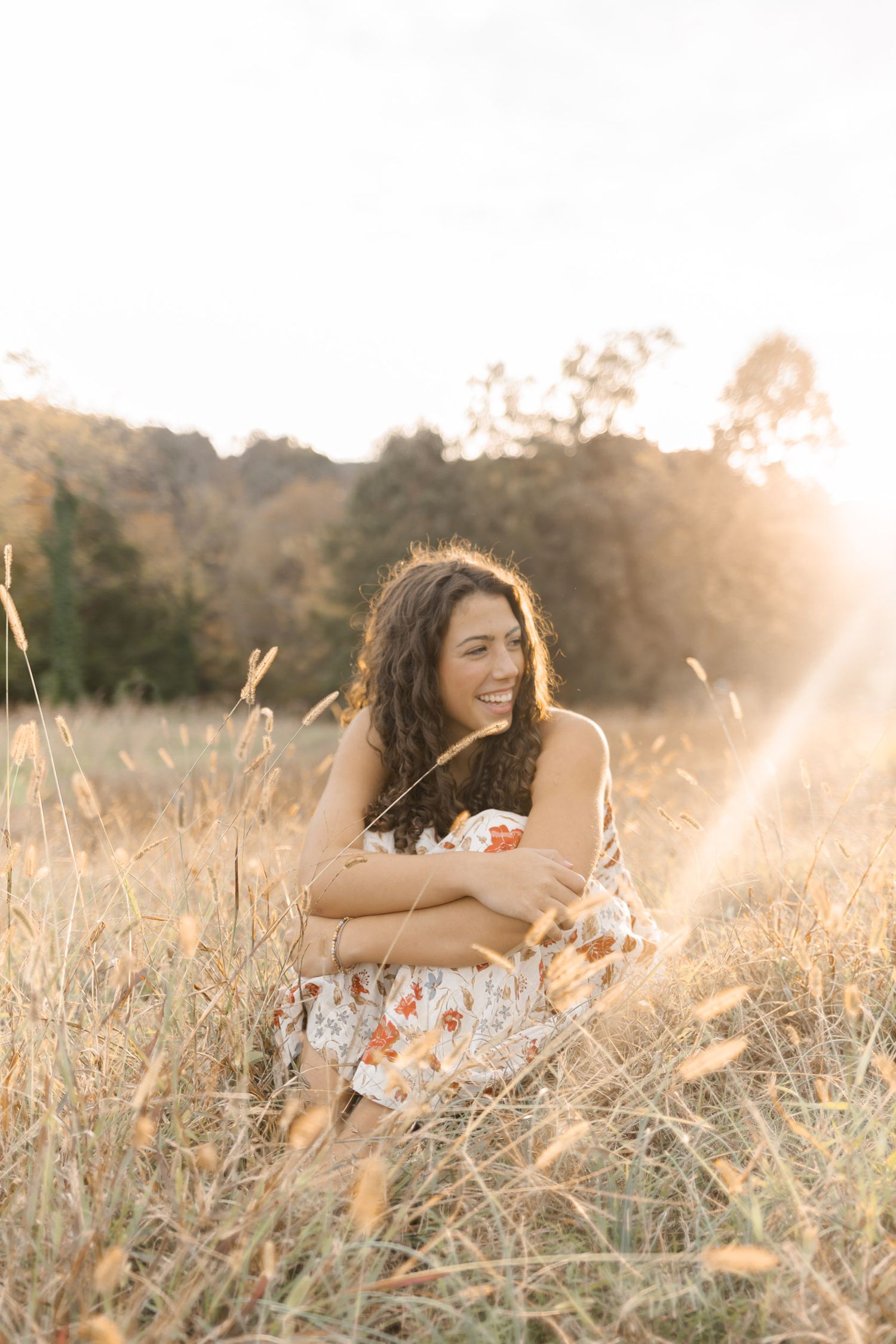 Young woman smiling joyfully while sitting in golden grass field during warm sunset light.