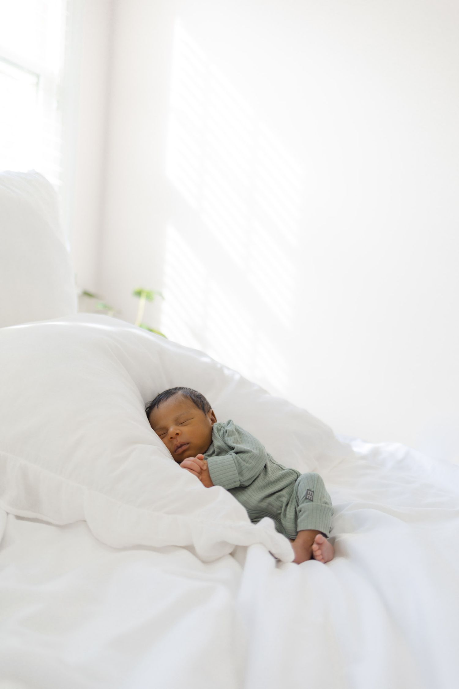 Newborn baby sleeping peacefully on white pillows in a bright room, wearing a green outfit.