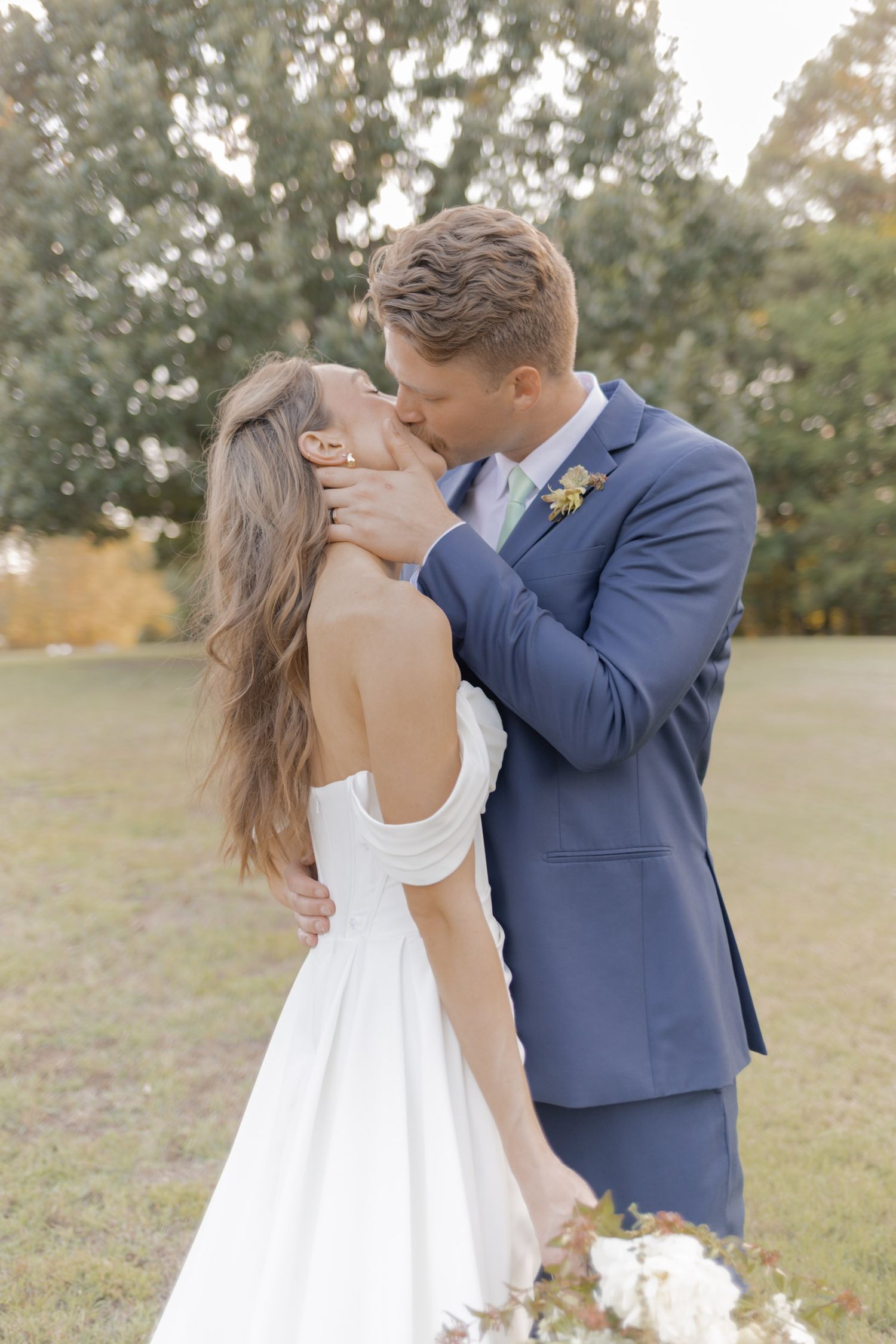A groom in a blue suit tenderly kisses his bride in a white gown outdoors during their wedding ceremony.