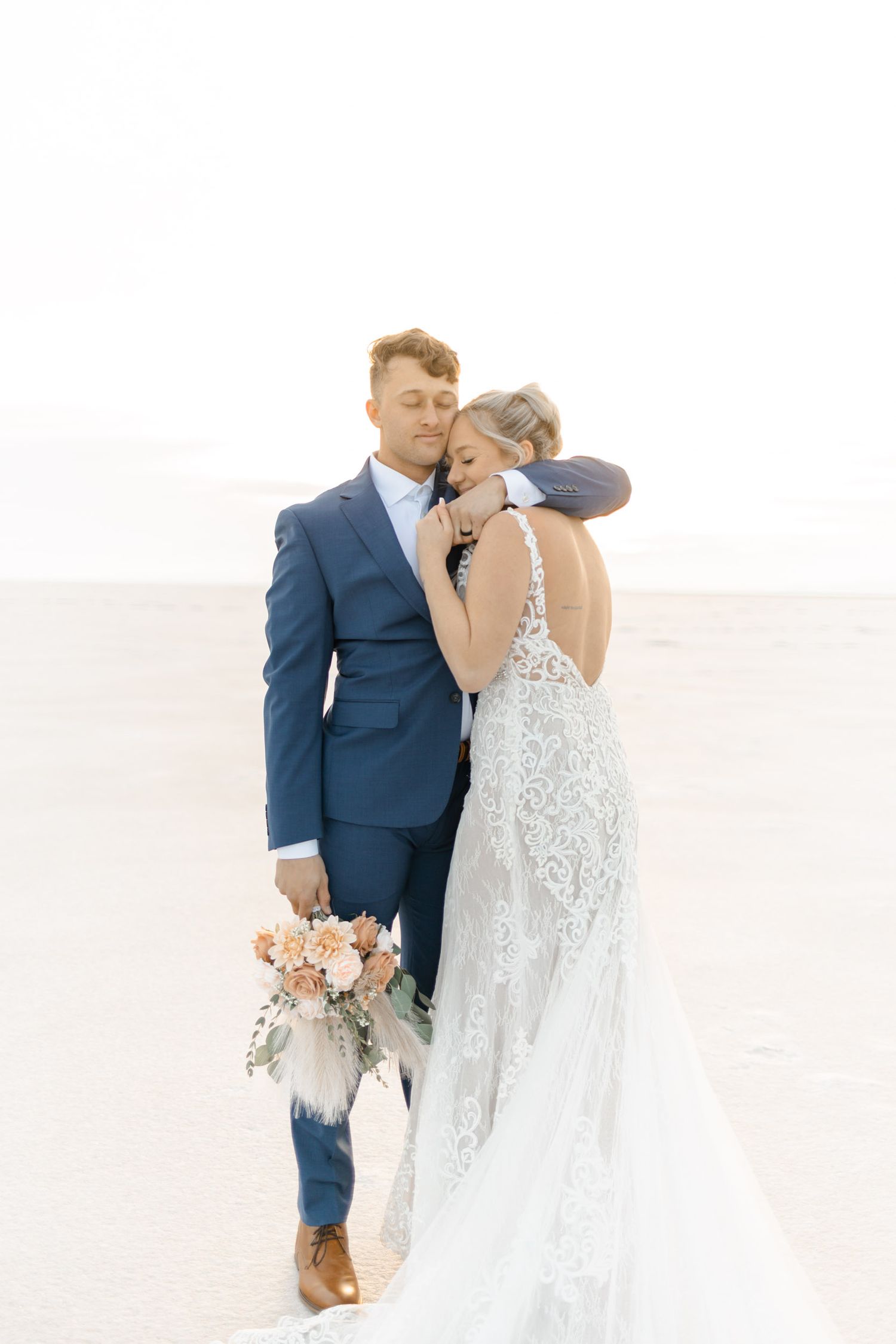 Romantic wedding couple embracing on white salt flats with soft peachy bouquet.