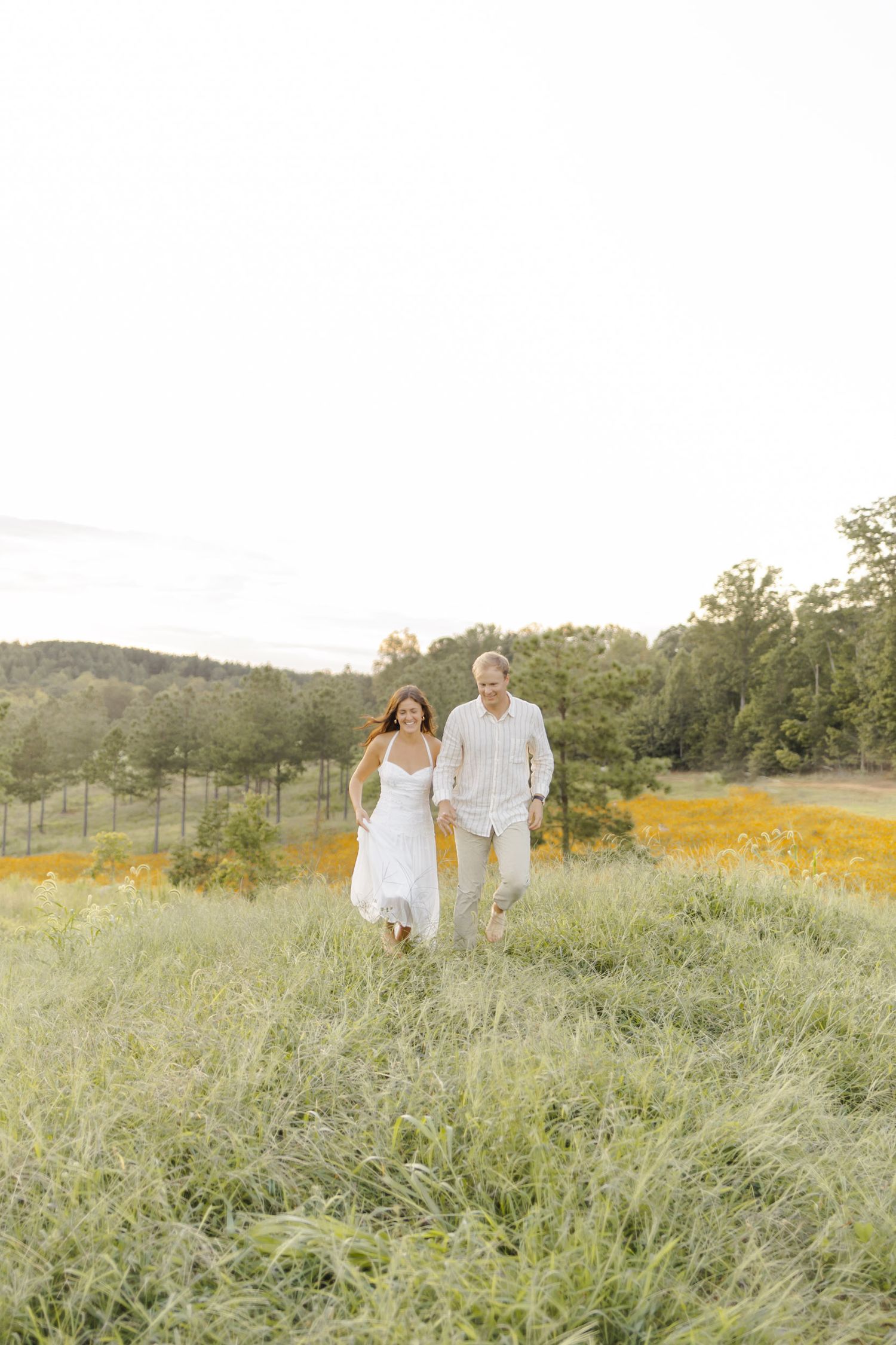 A couple in coordinated white outfits runs through a sun-drenched meadow during engagement session in Chapel Hill, NC.