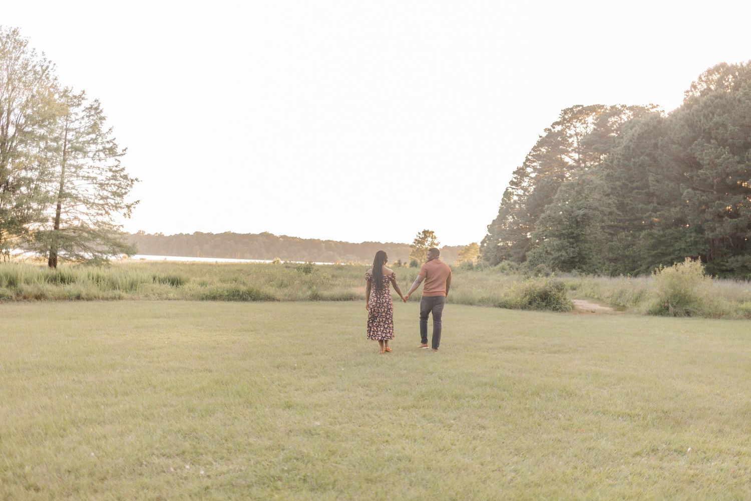 Couple holding hands walking through a lush green field at golden hour with trees and water in the background.