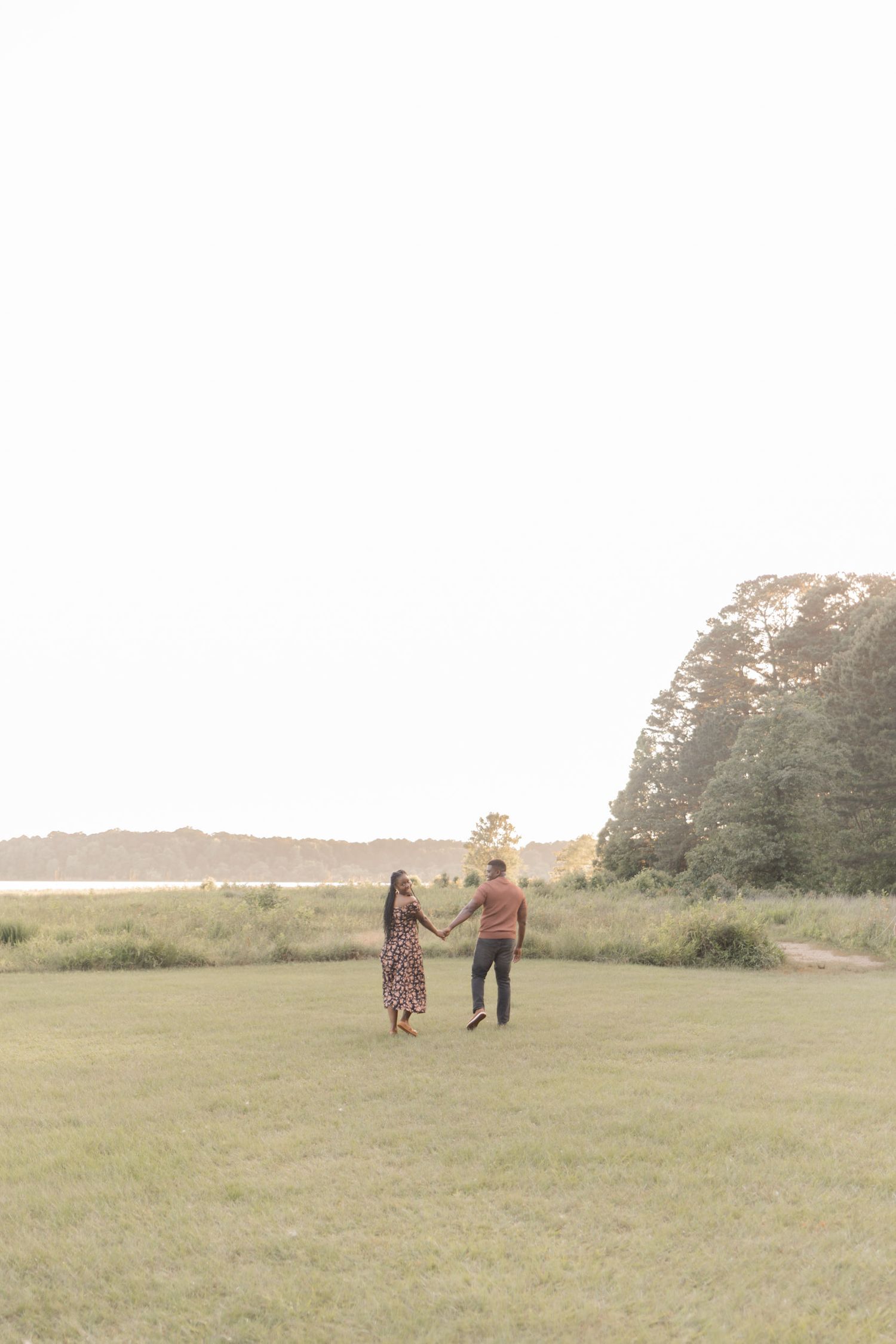 Couple holding hands and dancing in a green meadow near a serene lake surrounded by trees at golden hour.