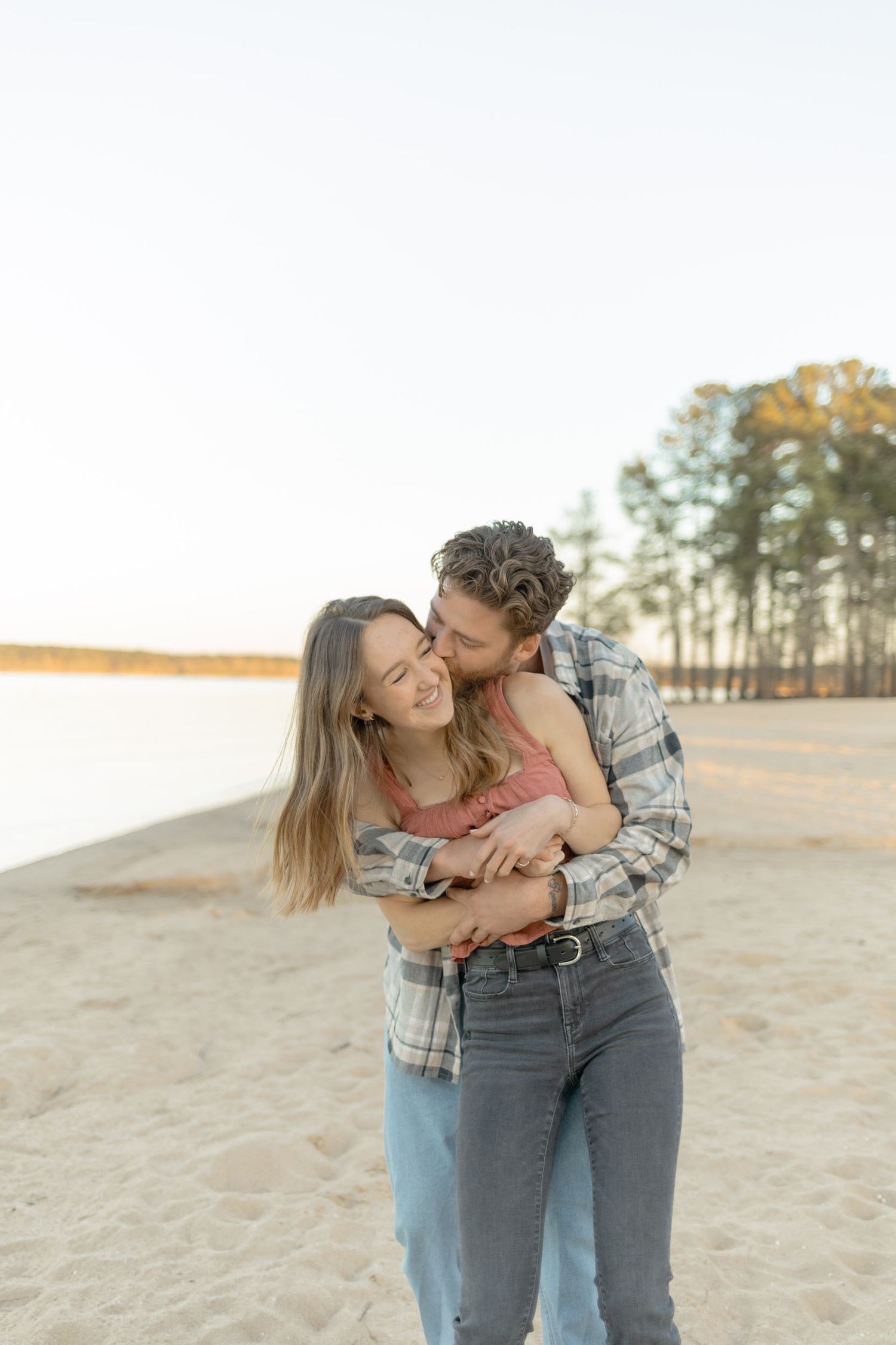 An intimate embrace captured on a sandy shoreline with pine trees and water in the background during their couples lifestyle photoshoot.