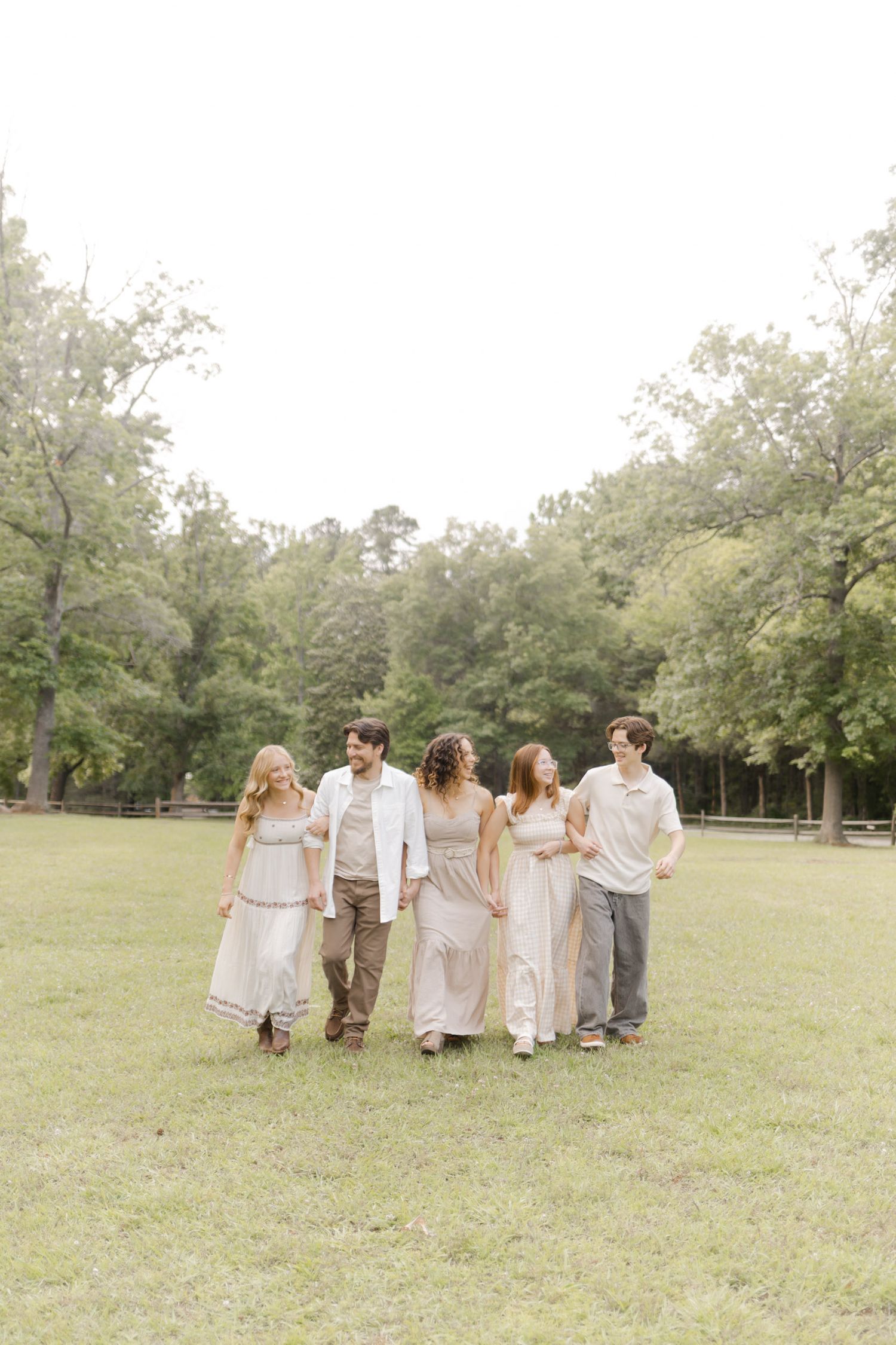 Friends in light colored dresses and grey pants walk arm in arm through a sunny park.