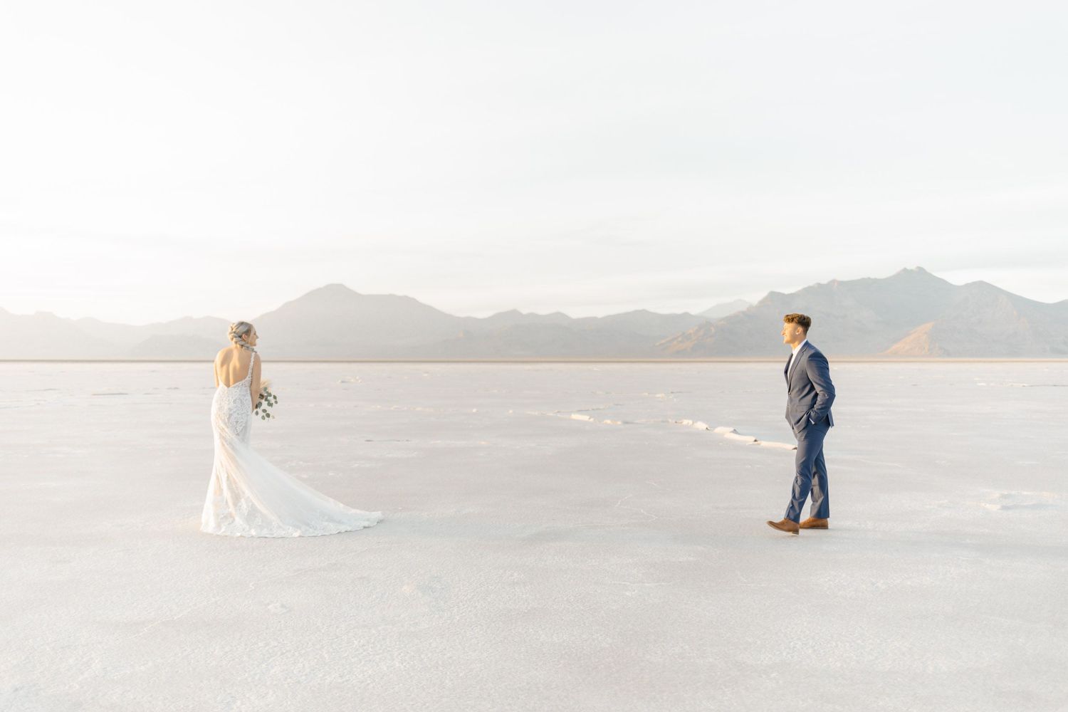 Two people in wedding attire stand apart on a vast white salt flat with mountains in the distance during sunset.