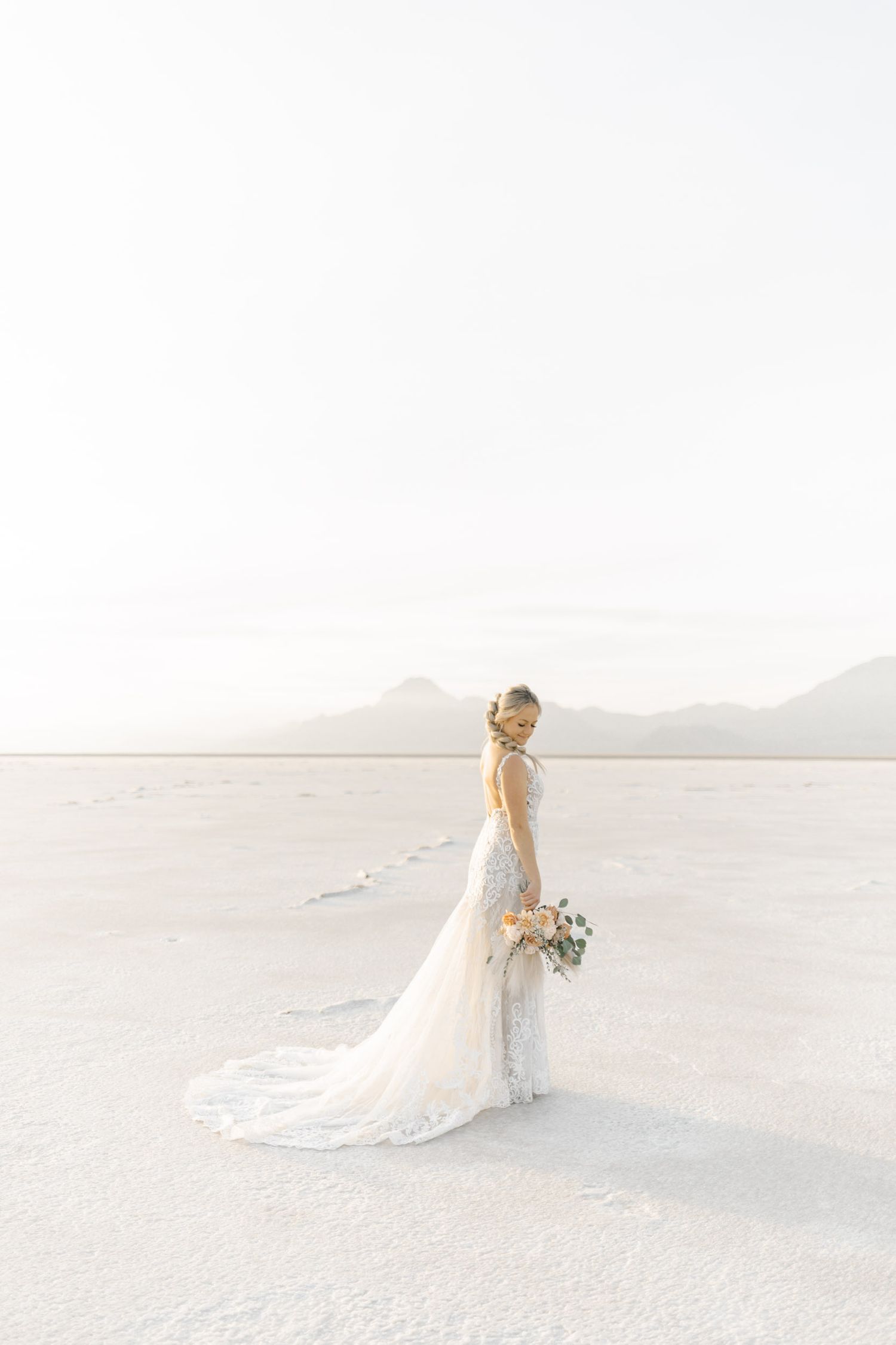 A bride in an elegant white dress poses against a minimalist landscape of salt flats and distant mountains.