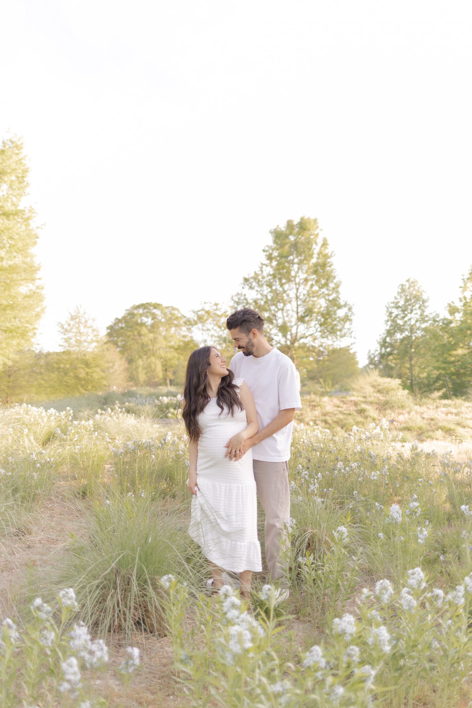 Expecting couple embracing in sunlit meadow with white wildflowers, woman in white maternity dress, golden hour lighting.