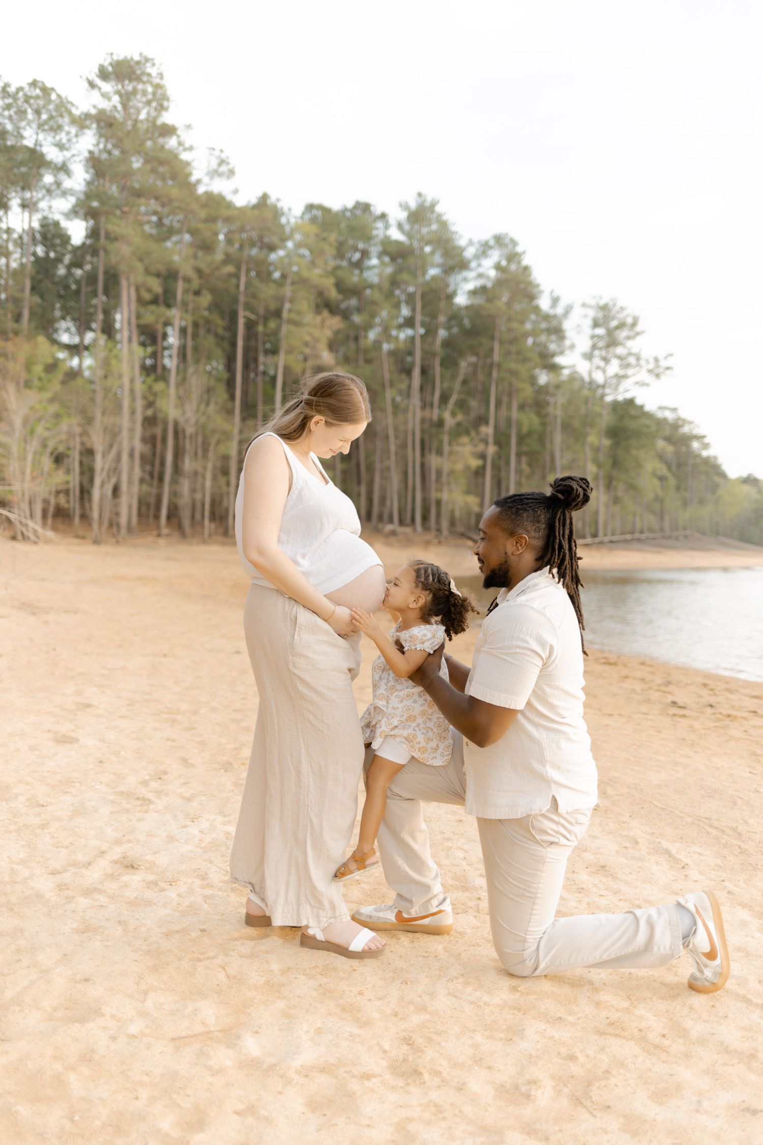 Interracial family maternity photo on sandy beach, father kneeling beside toddler kissing pregnant mothers belly.