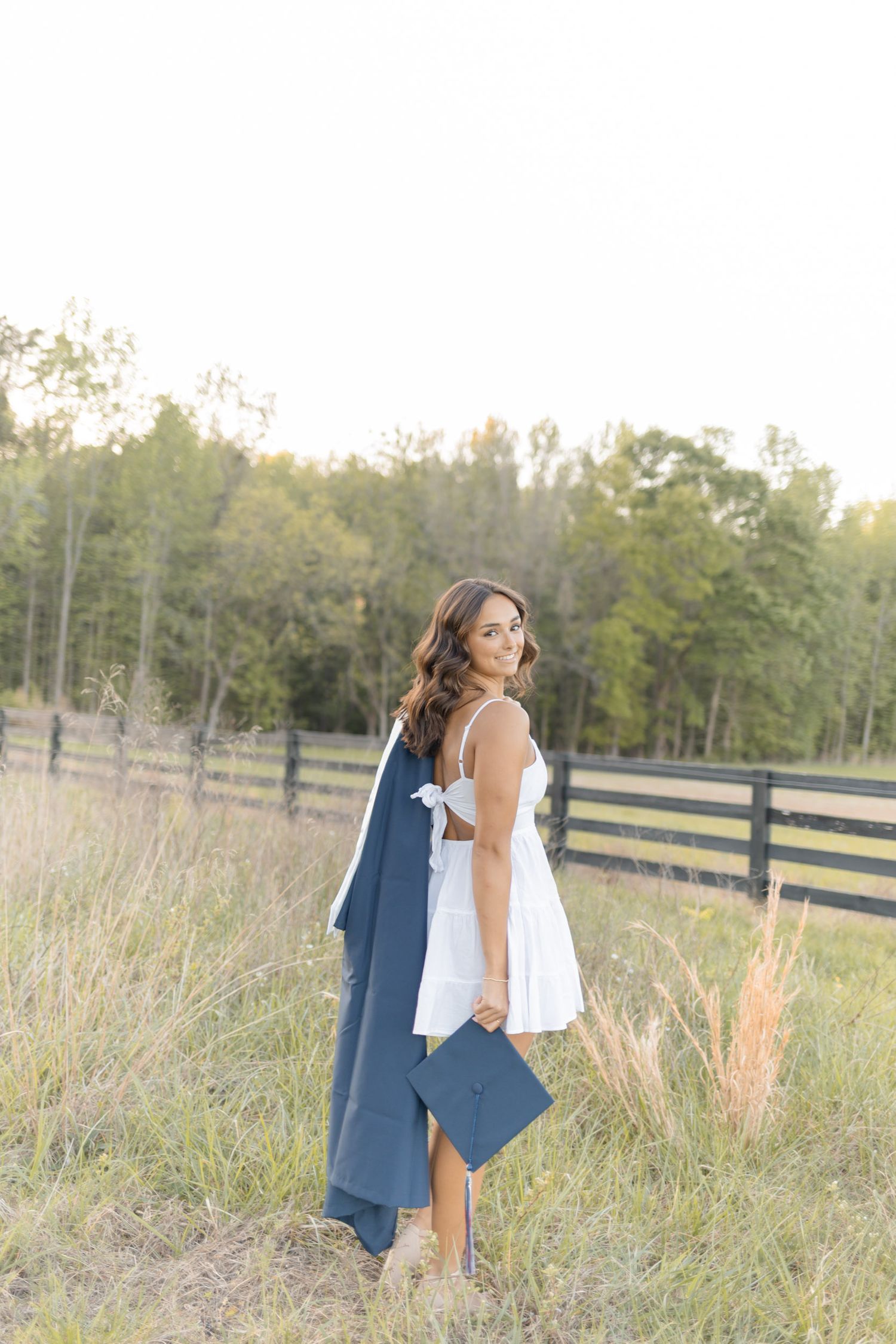 Young woman in white dress holds navy graduation gown and cap in a sunlit field with wooden fence and trees.