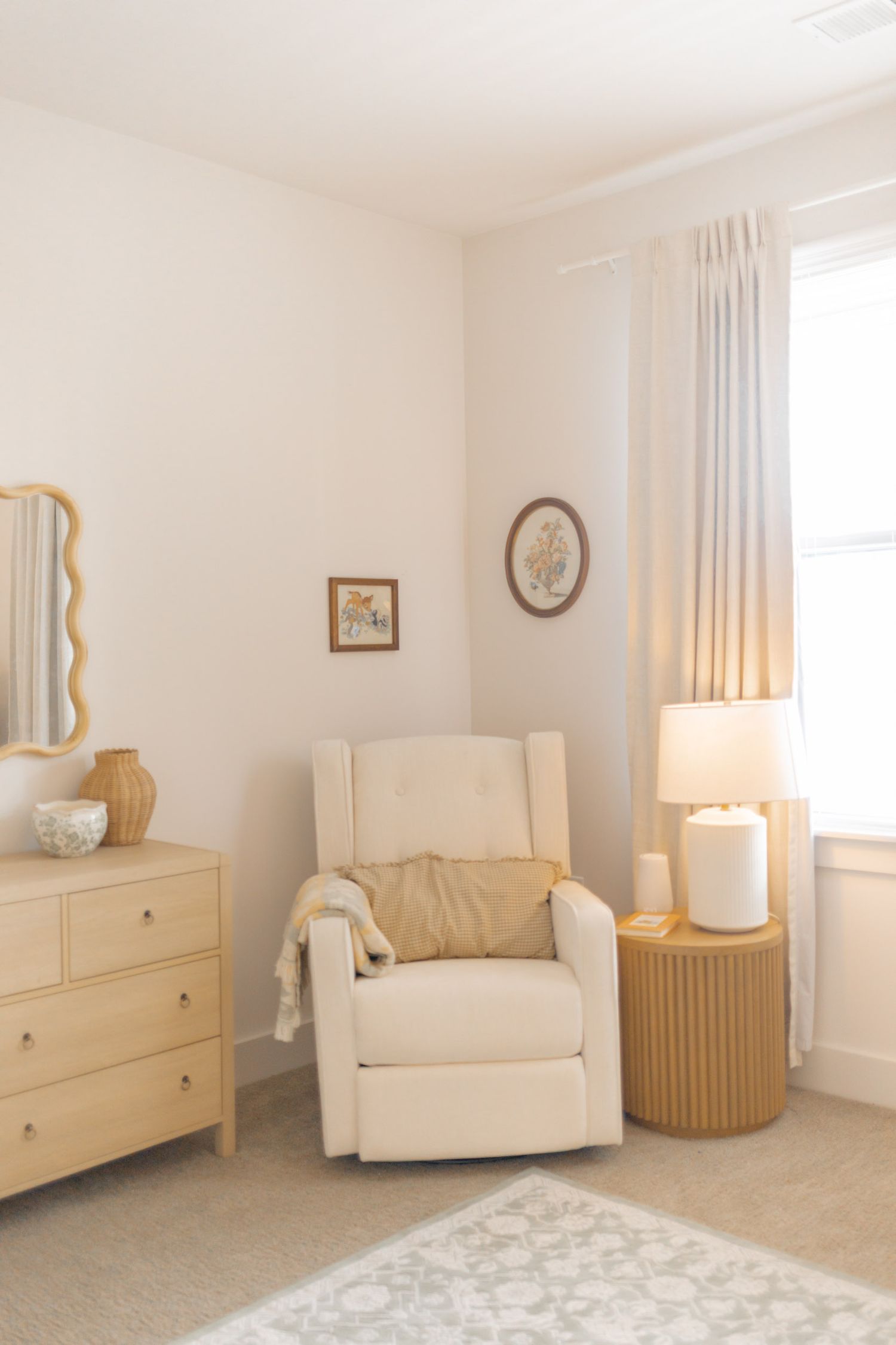 Neutral nursery corner with cream glider chair, wood dresser, wavy mirror, and soft lighting.
