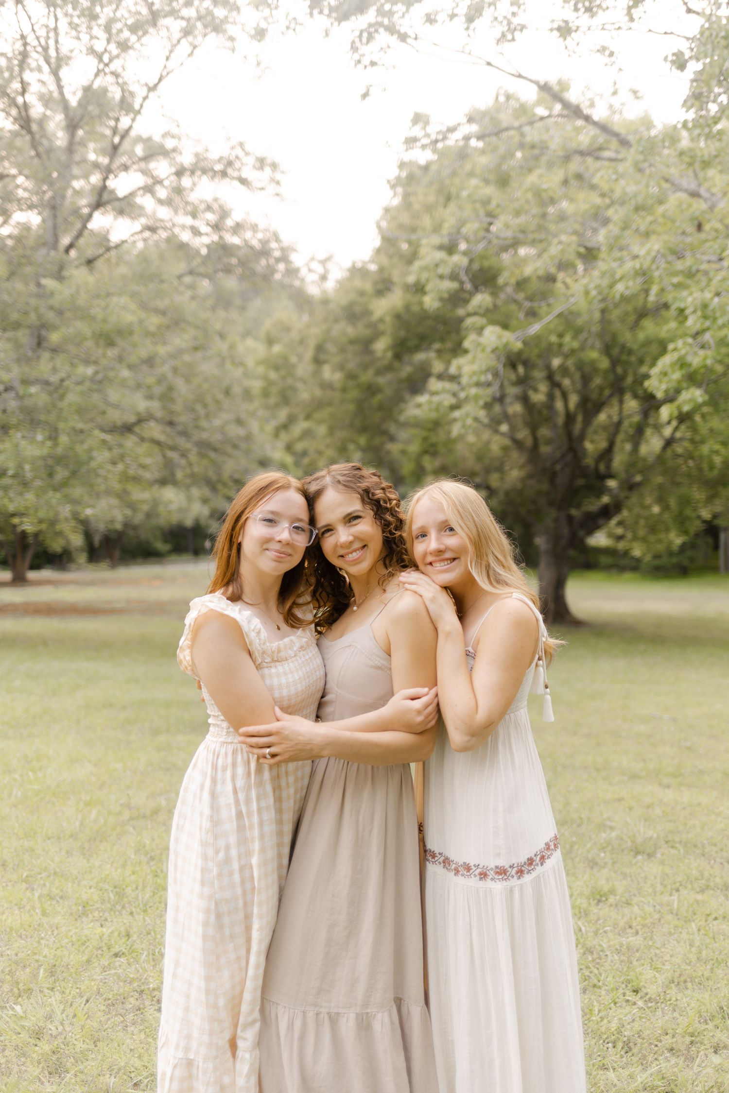 Three friends in light-colored dresses embrace while standing in a sunny field with trees in the background in Durham, North Carolina.
