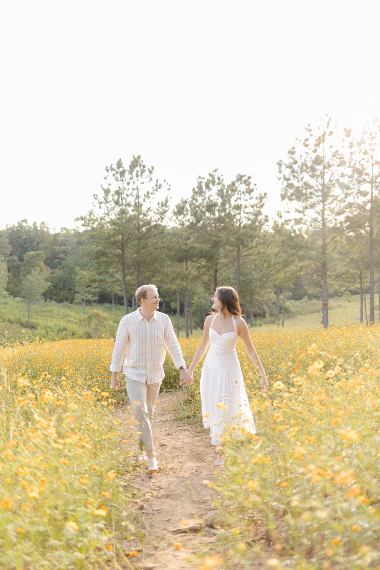 Couple walking hand in hand through a path surrounded by yellow wildflowers and pine trees during lifestyle portrait photoshoot.