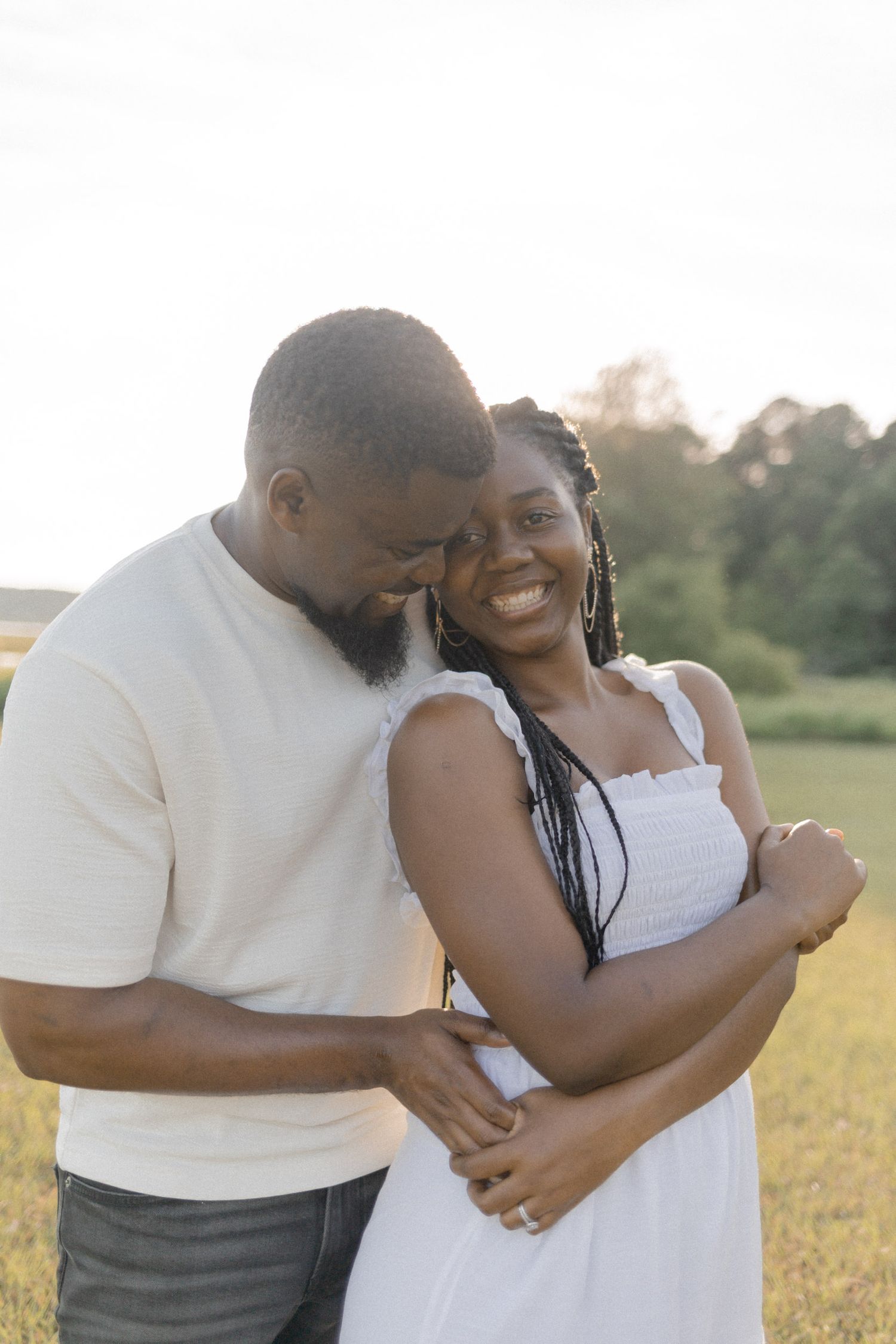 A joyful couple embraces in a sunlit field, the woman smiling warmly as the man nuzzles her head.