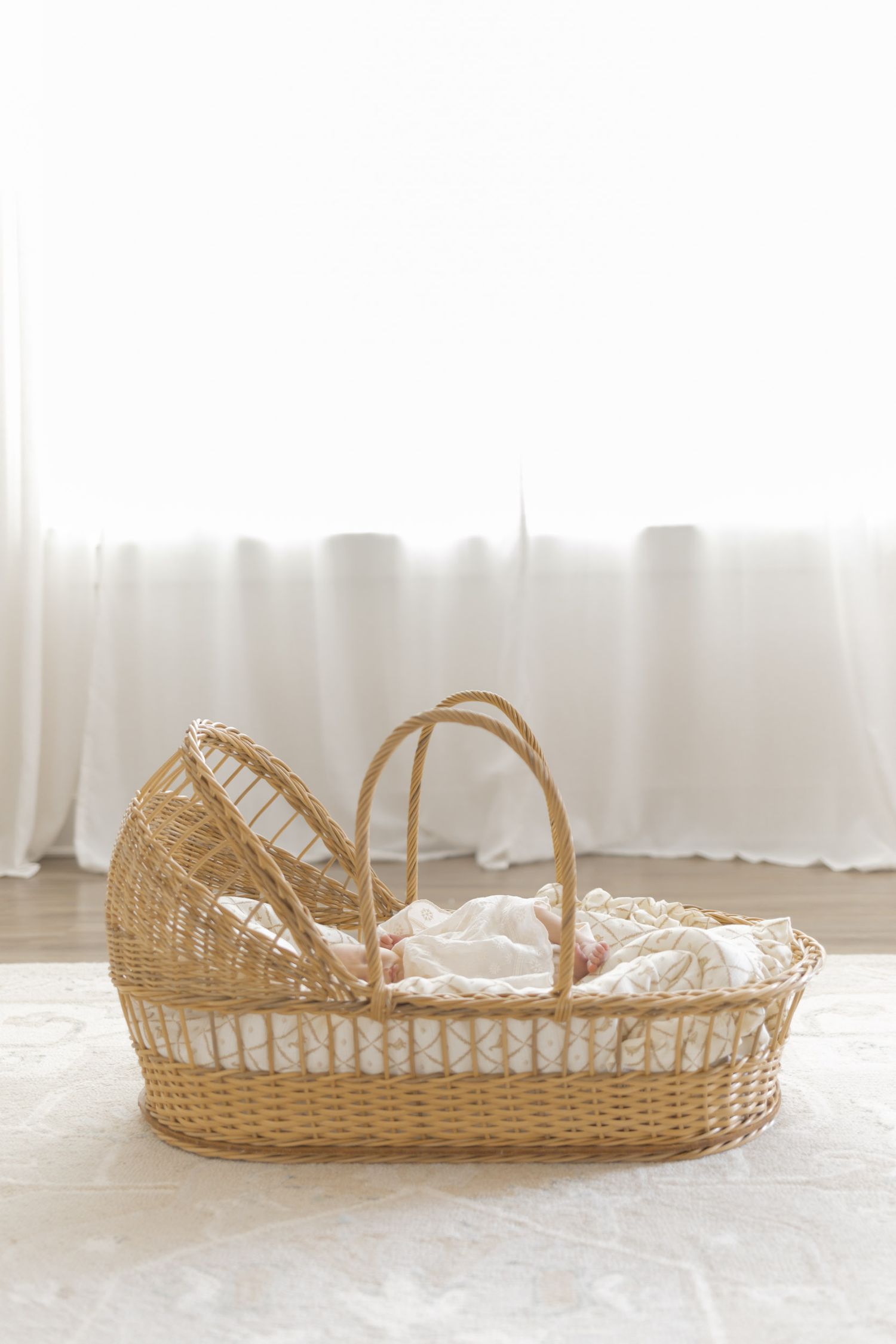 Wicker Moses basket with white linen bedding placed on a rug near a bright window.