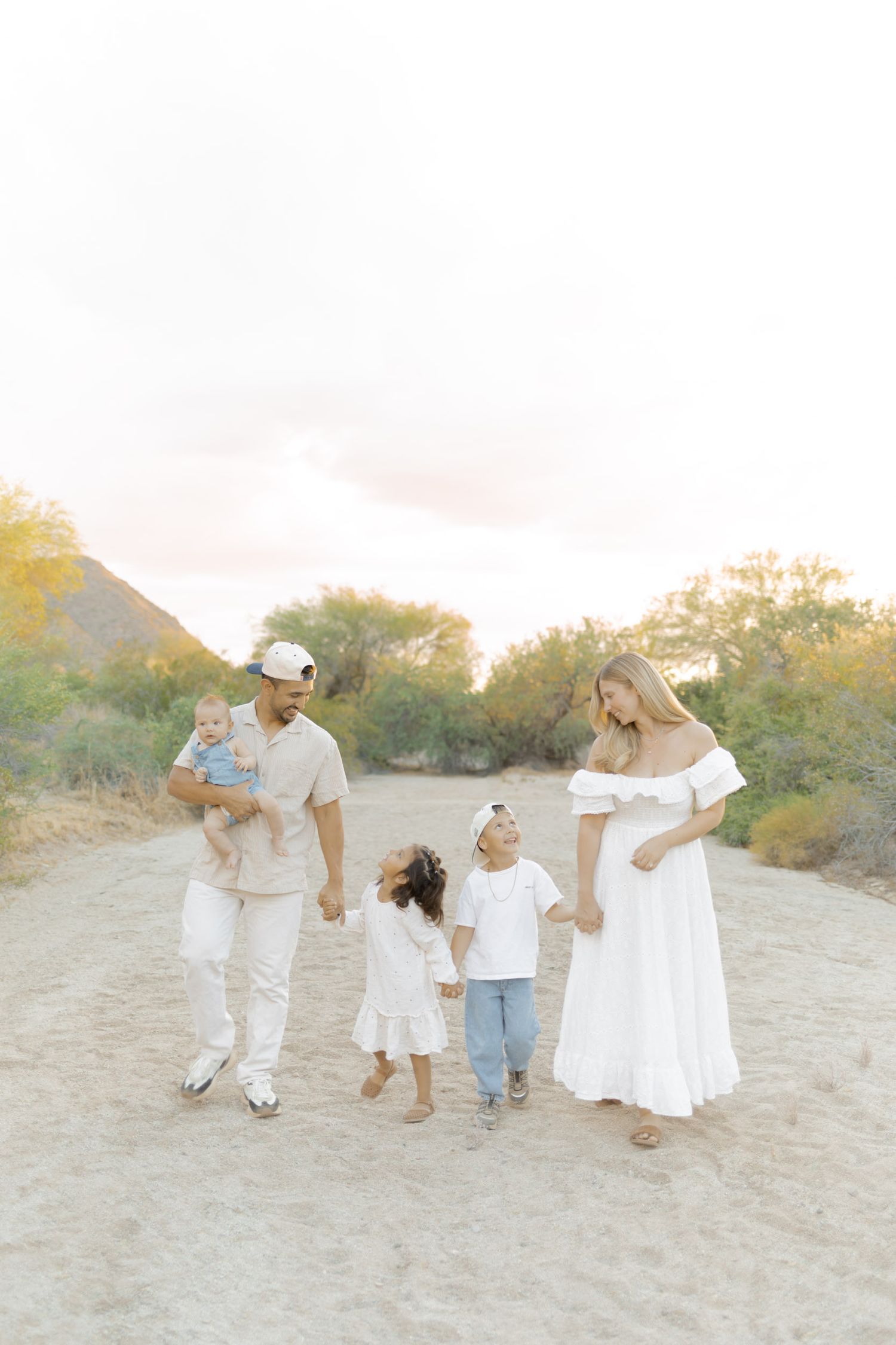 Family of five walking on sandy desert path, parents with three young children dressed in white and neutral tones at golden hour.