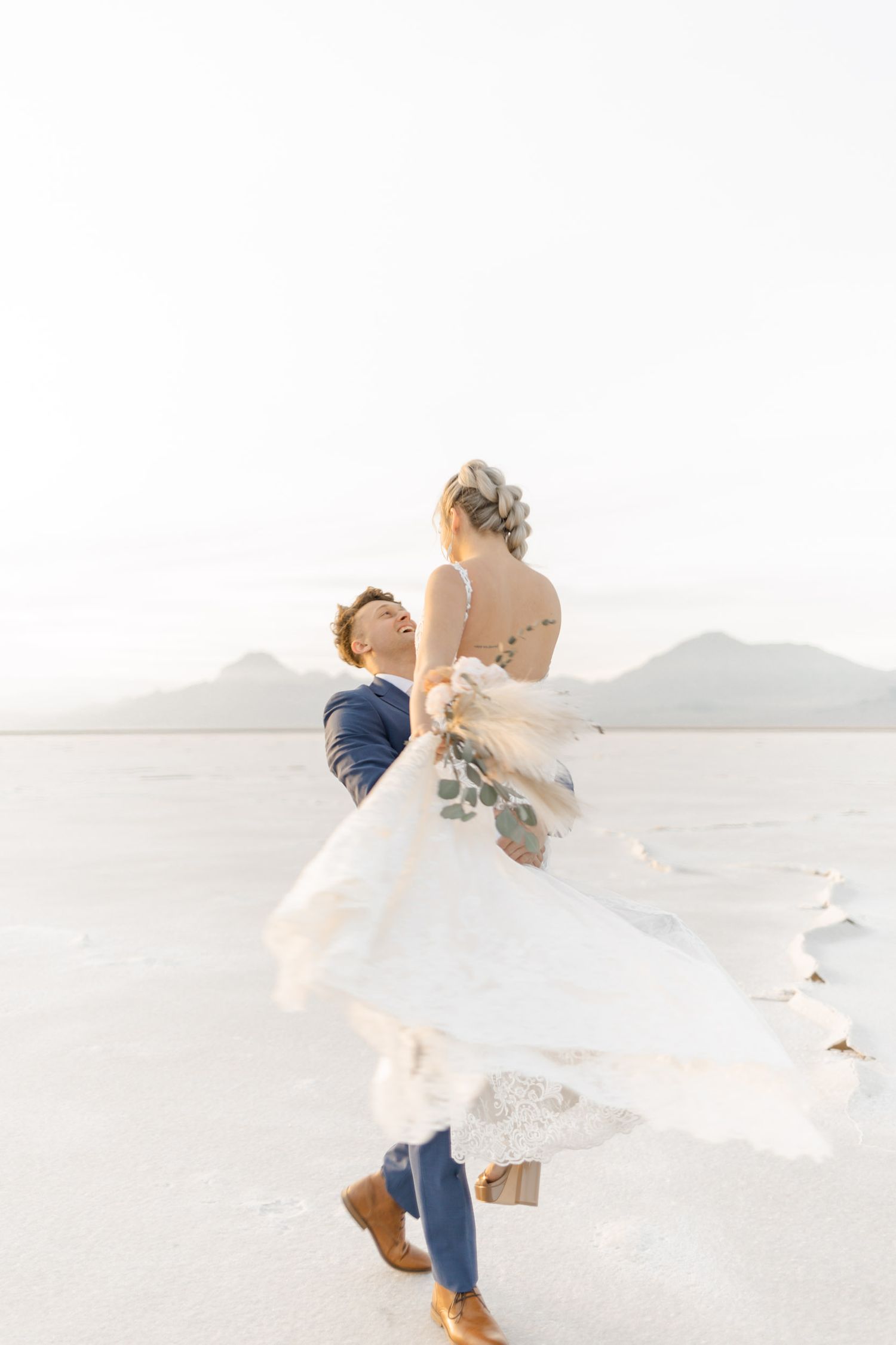 Newlyweds dance and twirl joyfully on a pristine white beach with mountains in the background.