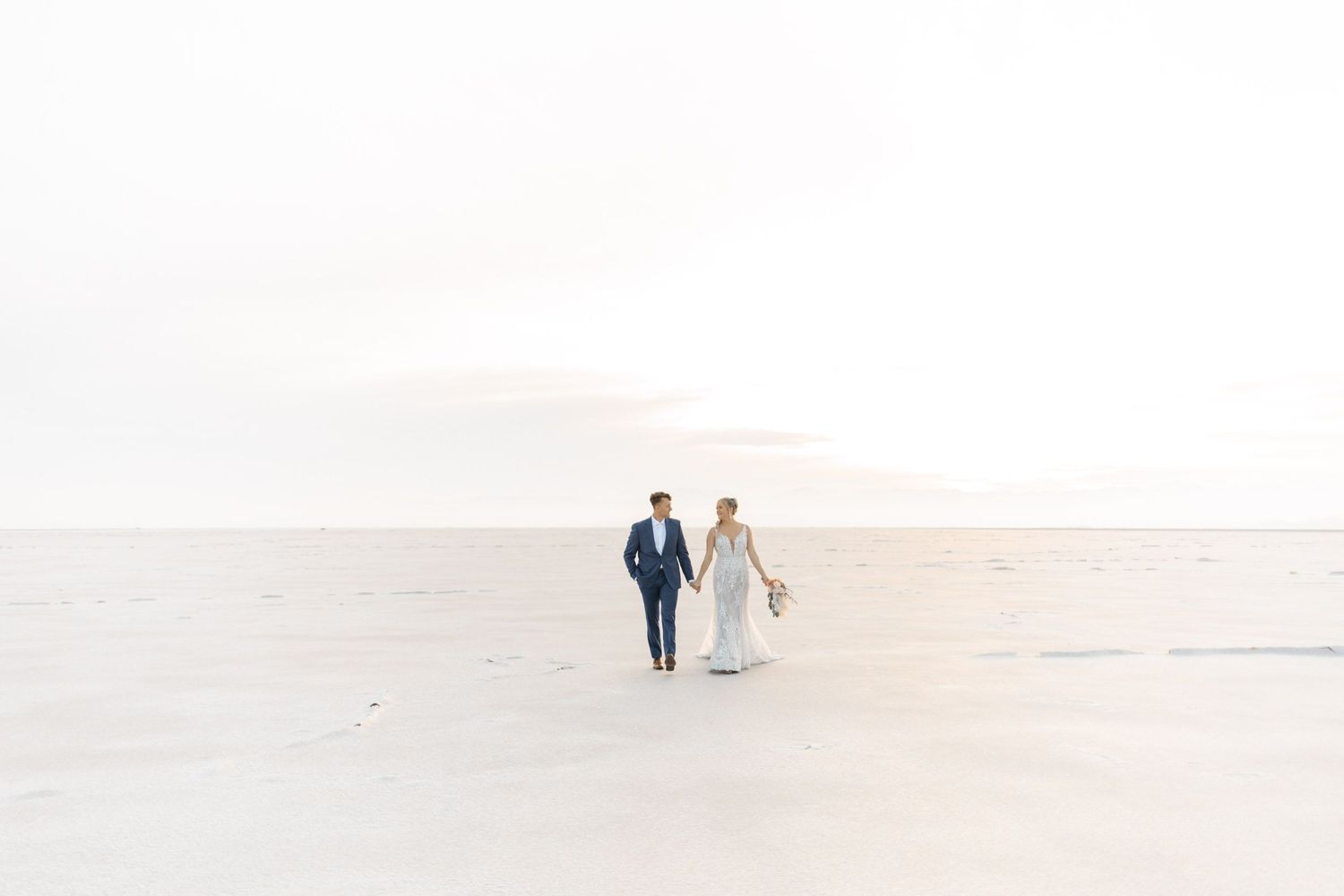 A couple walks hand in hand along a pristine white beach with soft waves lapping at the shore.