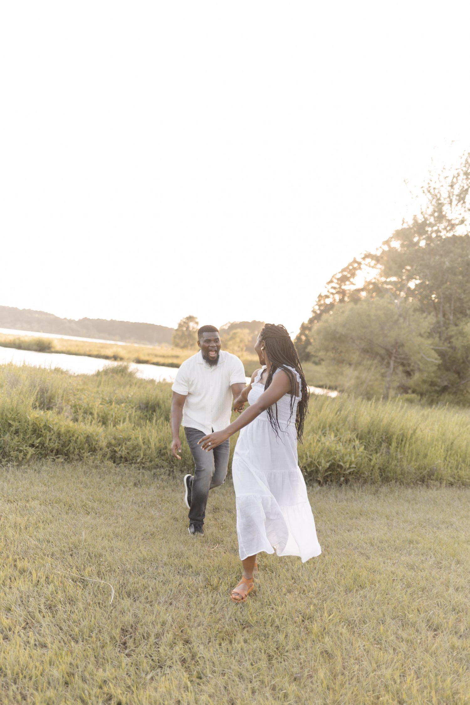 A joyful Black couple holds hands while walking through a sunlit green field at golden hour, woman in white dress.