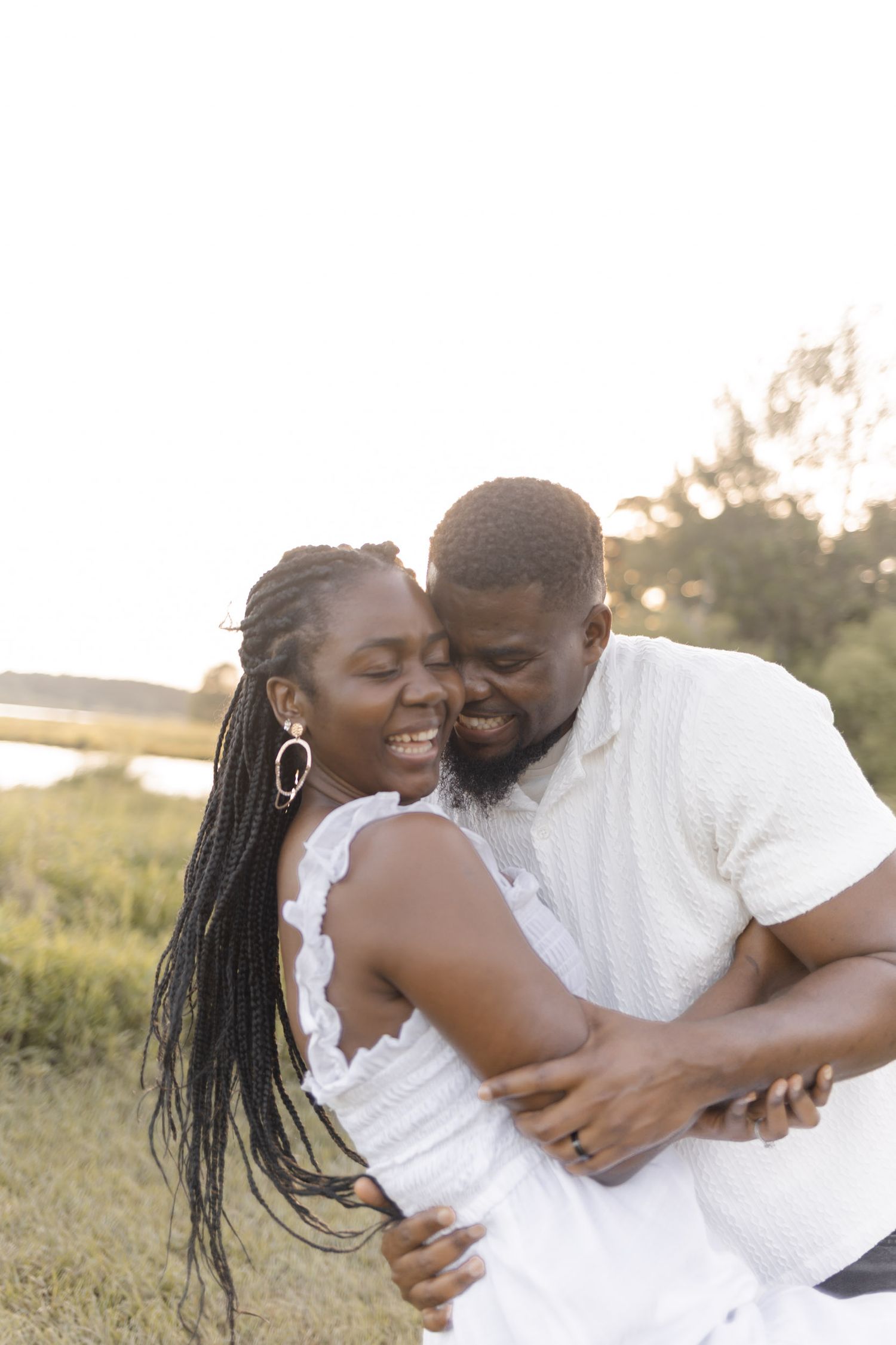 A joyful Black couple embraces outdoors at golden hour, both wearing white, sharing a tender romantic moment in a grassy field.