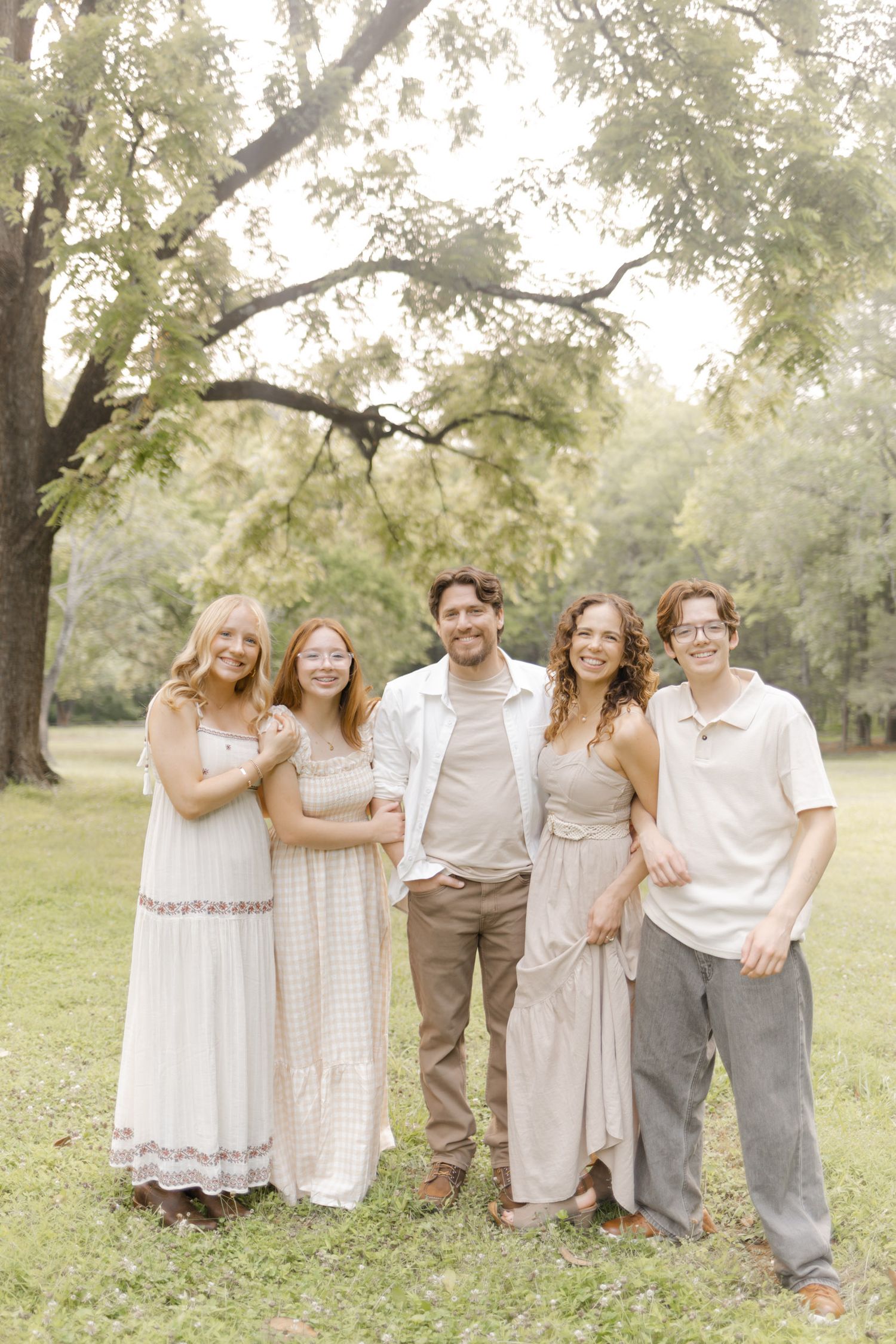 Group portrait of people in coordinated beige and white outfits standing together under large trees while doing a photoshoot.