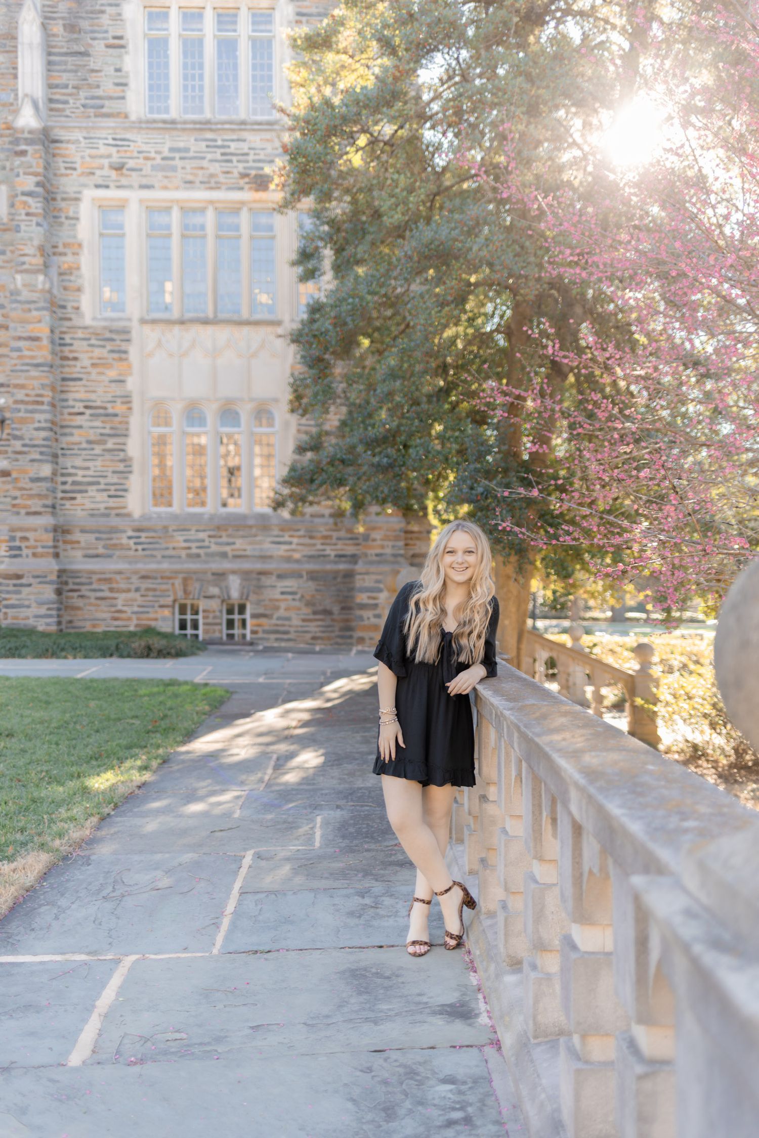 Young woman in black dress posing by stone wall near historic building with blooming trees and sunlight.