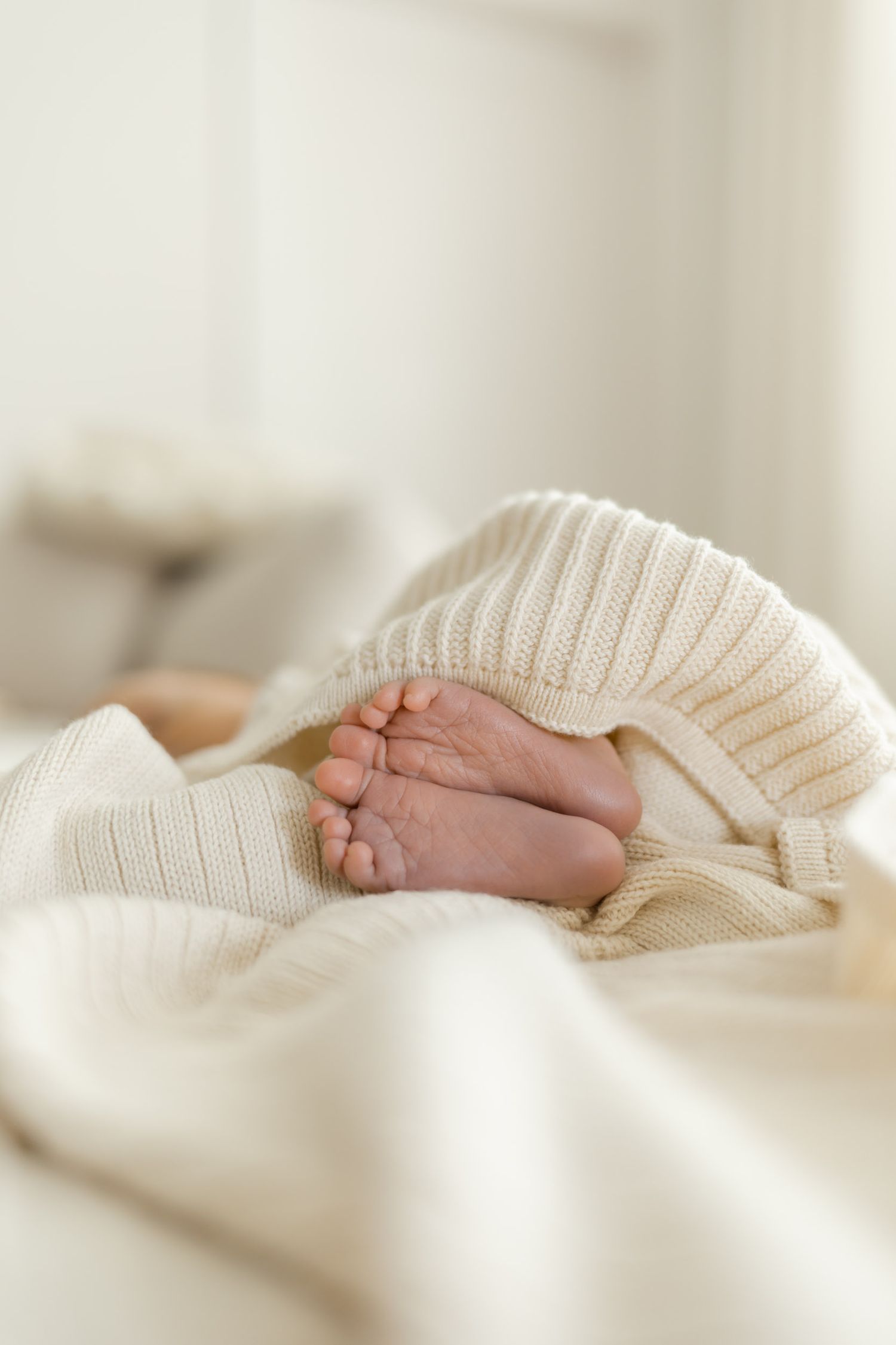 Newborn baby feet peeking out from a cozy cream knit blanket on a soft white bed.