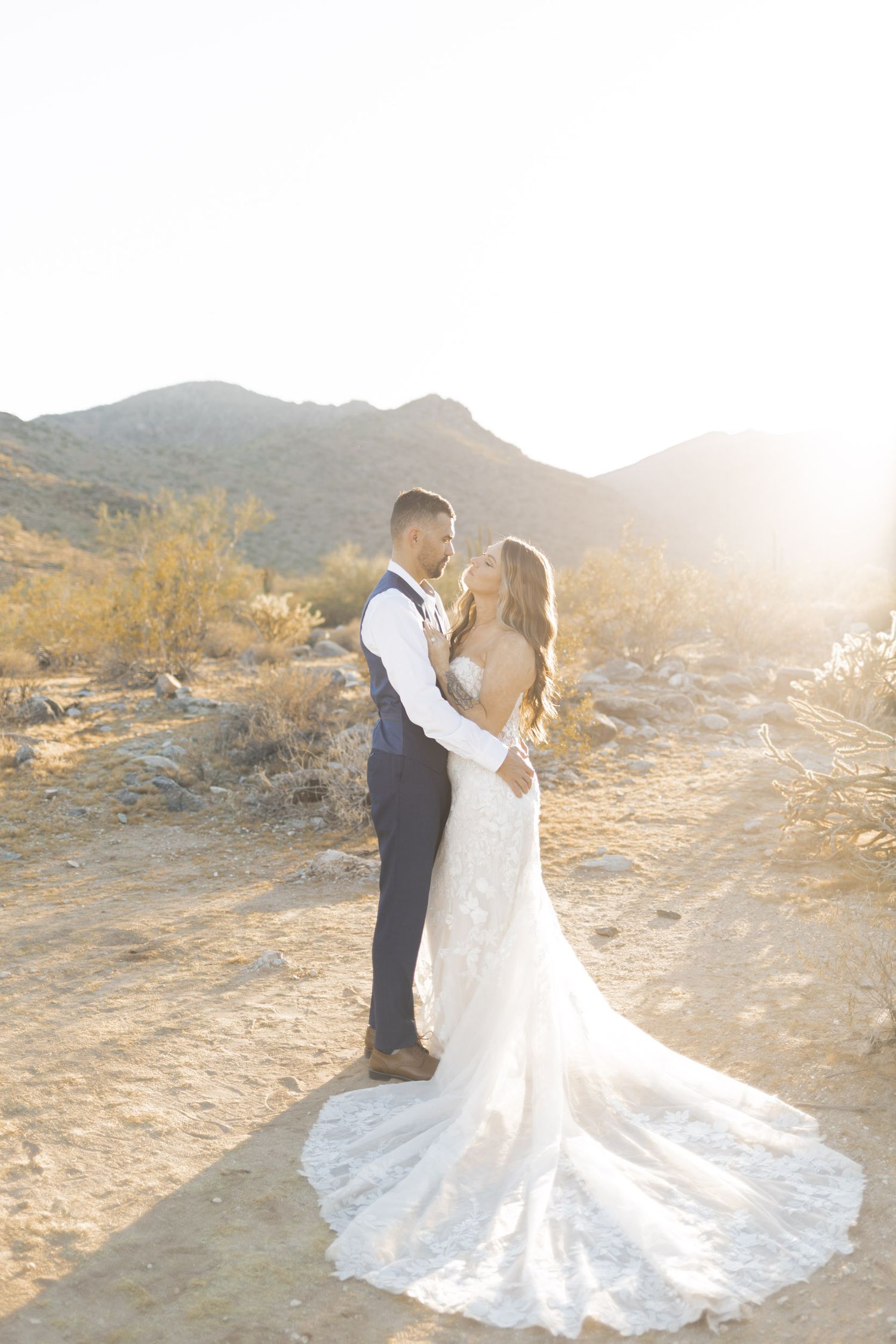 Bride and groom share a romantic moment in a sunlit desert landscape with mountains, bride in lace gown with long train.