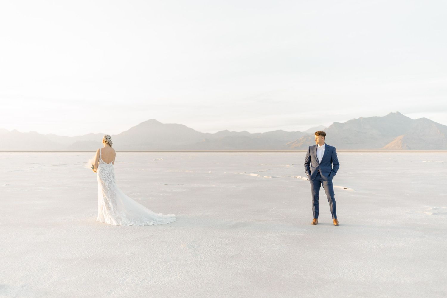 A couple in wedding attire walks along a scenic beach with mountains in the distance at sunset.