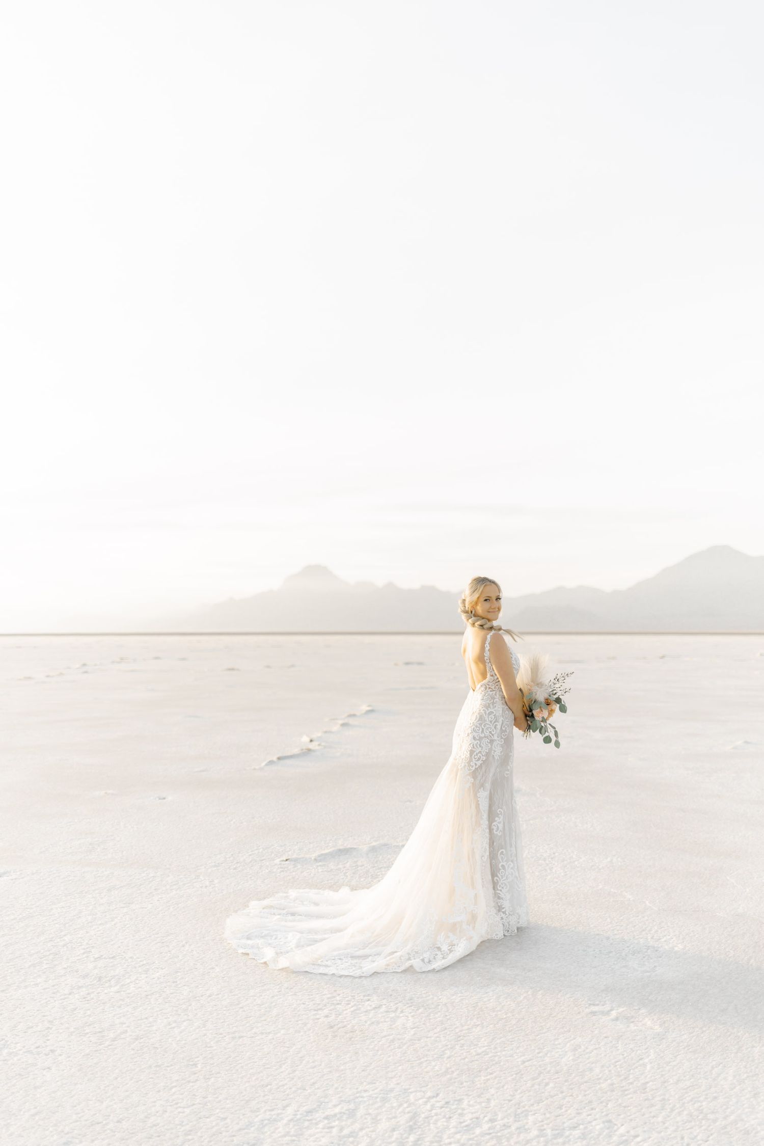 A bride in a flowing white lace gown stands alone on a stark white salt flat with mountains behind her.