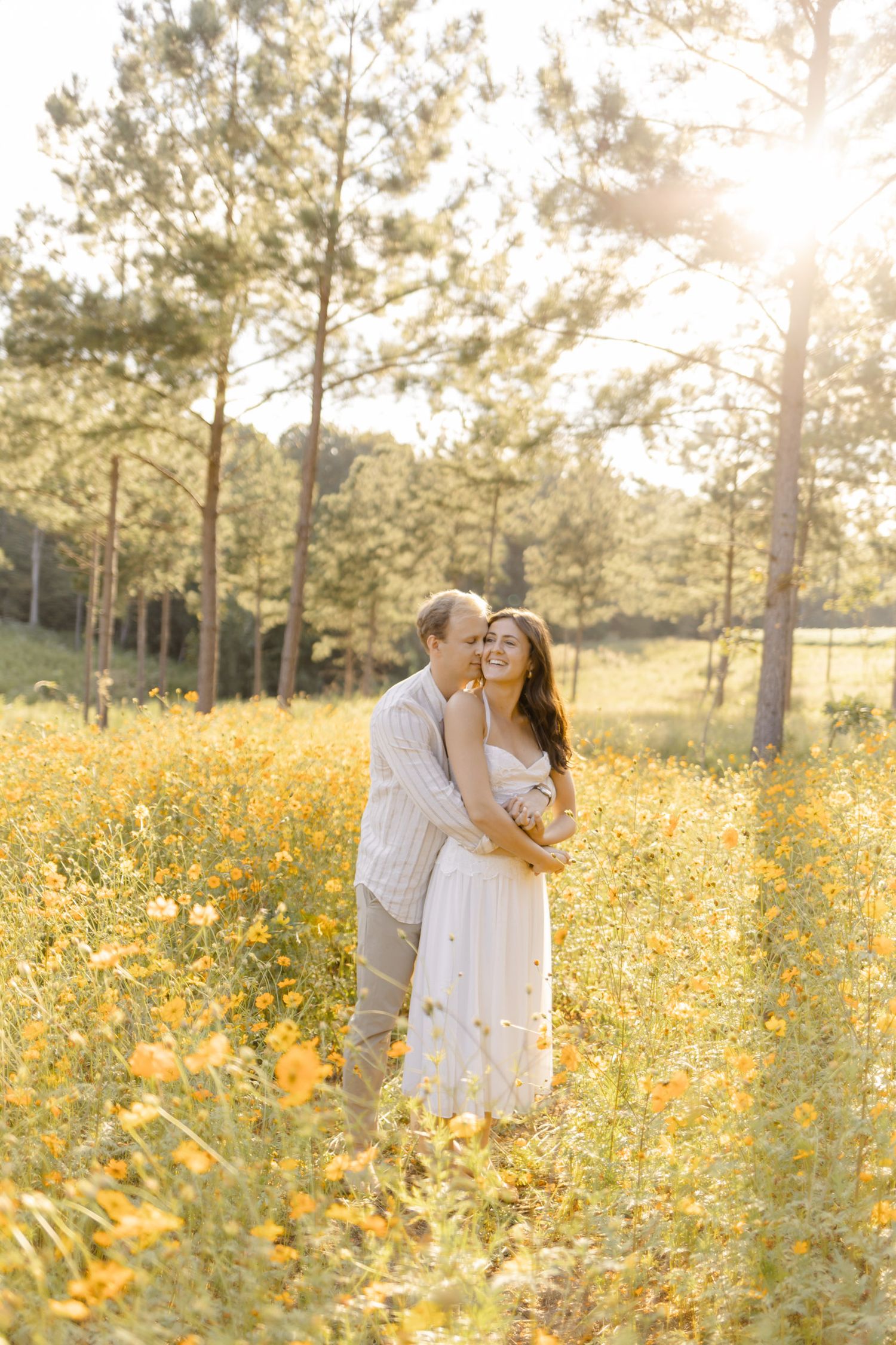 A romantic couple embraces in a sunlit field of yellow wildflowers with pine trees in the background for engagement session in Chapel Hill.