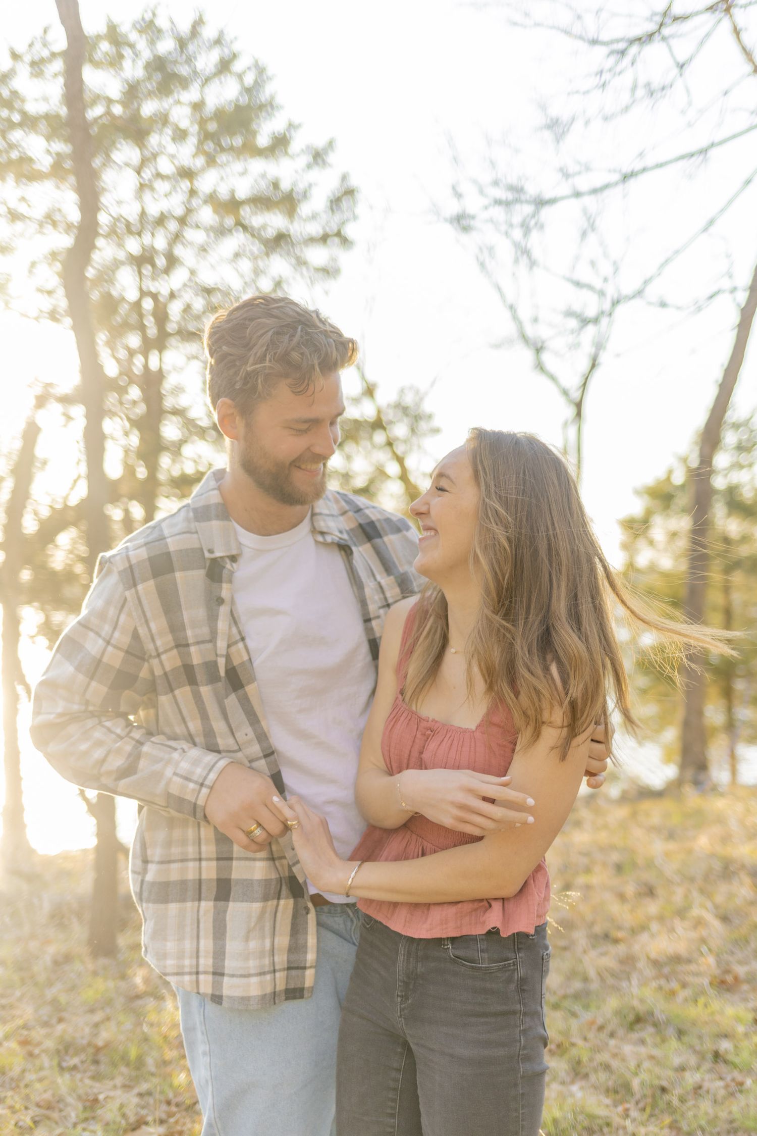 Couple laughing and embracing in casual attire against a backdrop of pine trees.