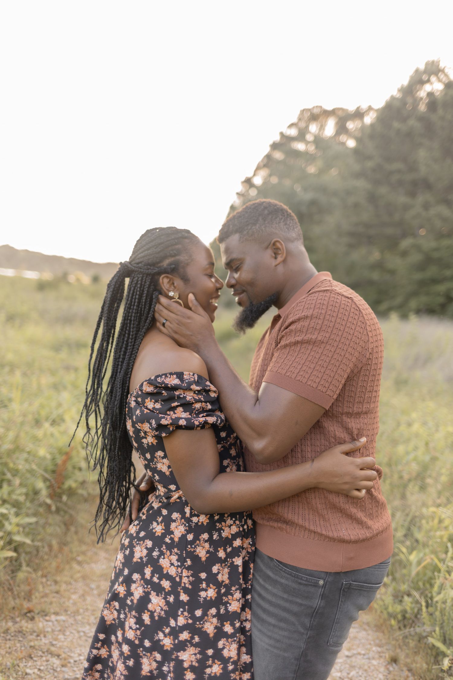 A romantic Black couple nearly kisses while embracing on a nature path at sunset, she in floral dress, he in rust shirt.