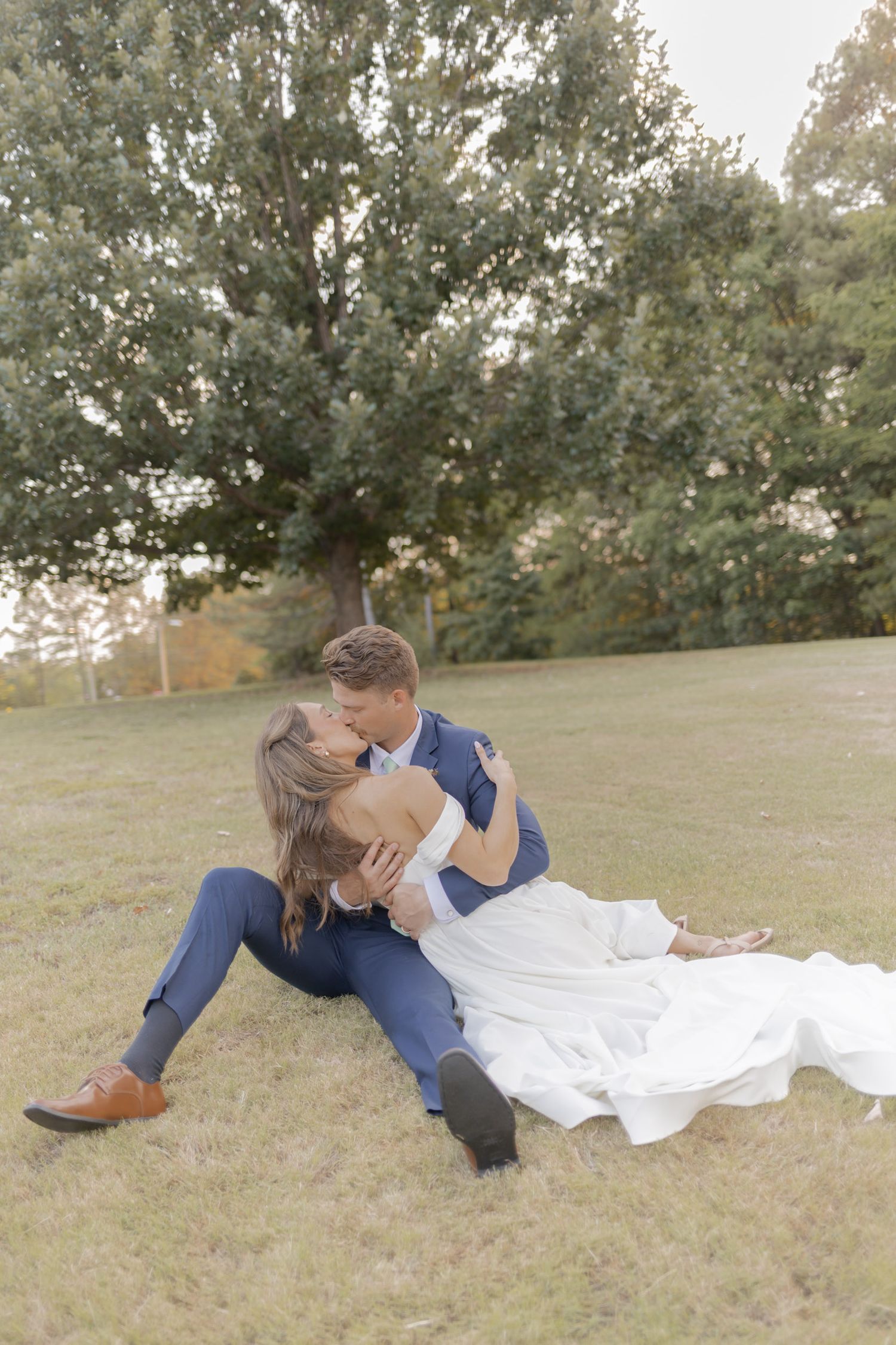 Bride and groom sitting on grass kissing, groom in blue suit, bride in white off-shoulder gown, trees in background.