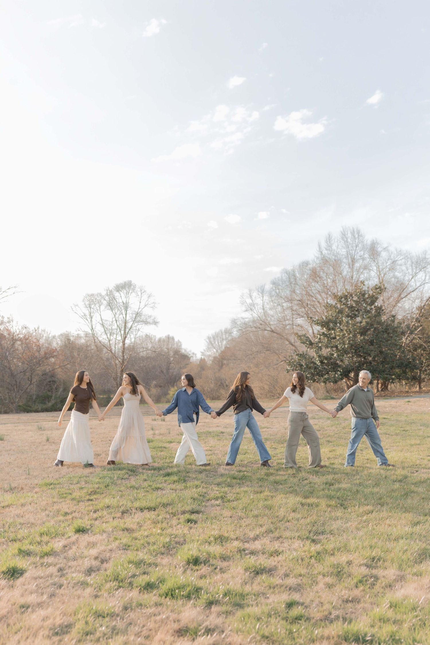 Six people hold hands in a line walking through a grassy field with bare trees in the background.