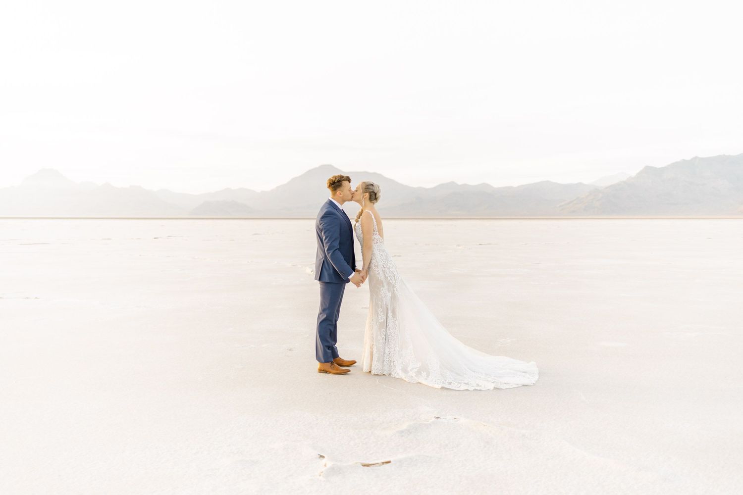 A romantic wedding couple embraces on a scenic white beach with gentle waves in the background.