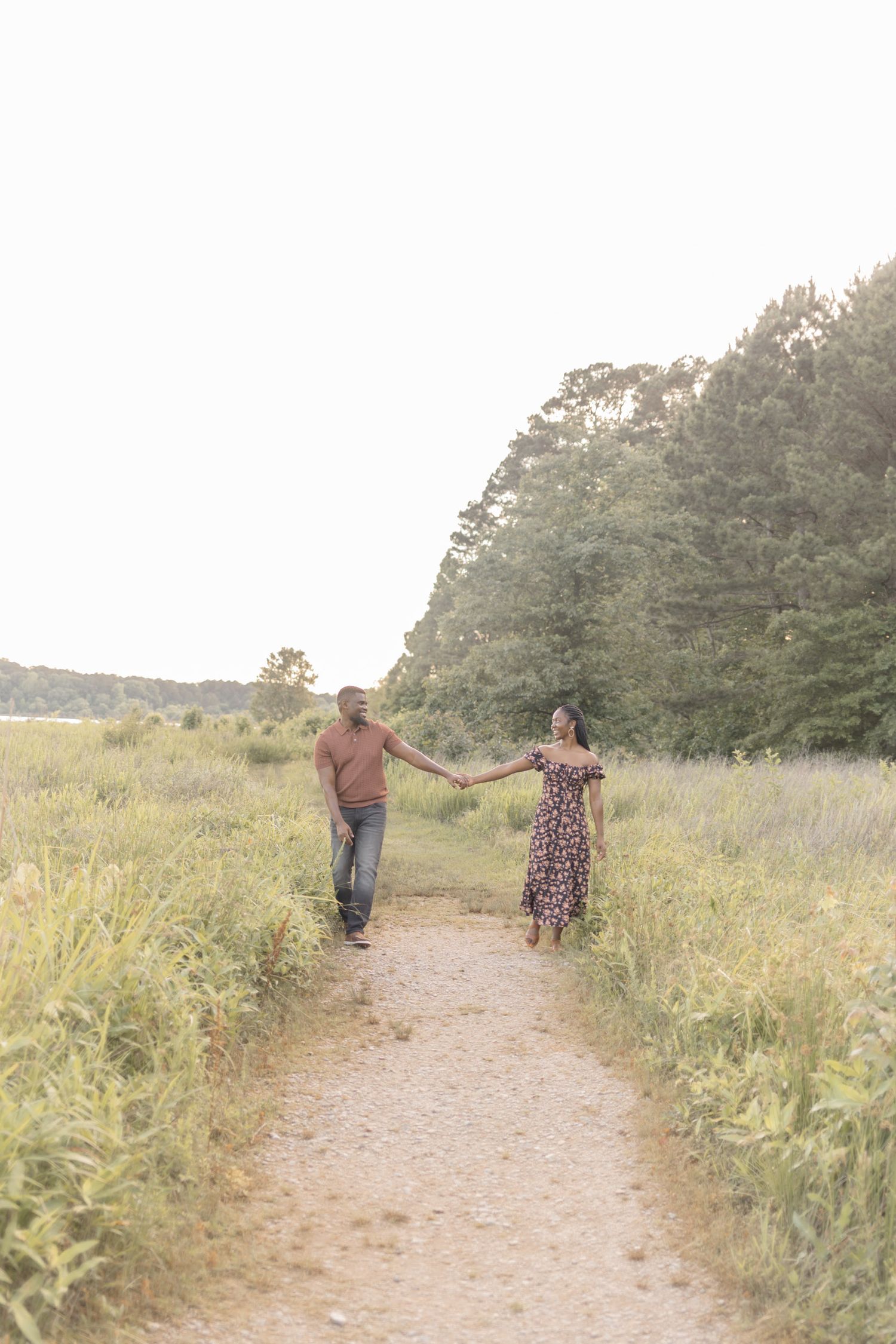 Couple holding hands walking on a scenic nature path surrounded by lush green fields and trees at golden hour.