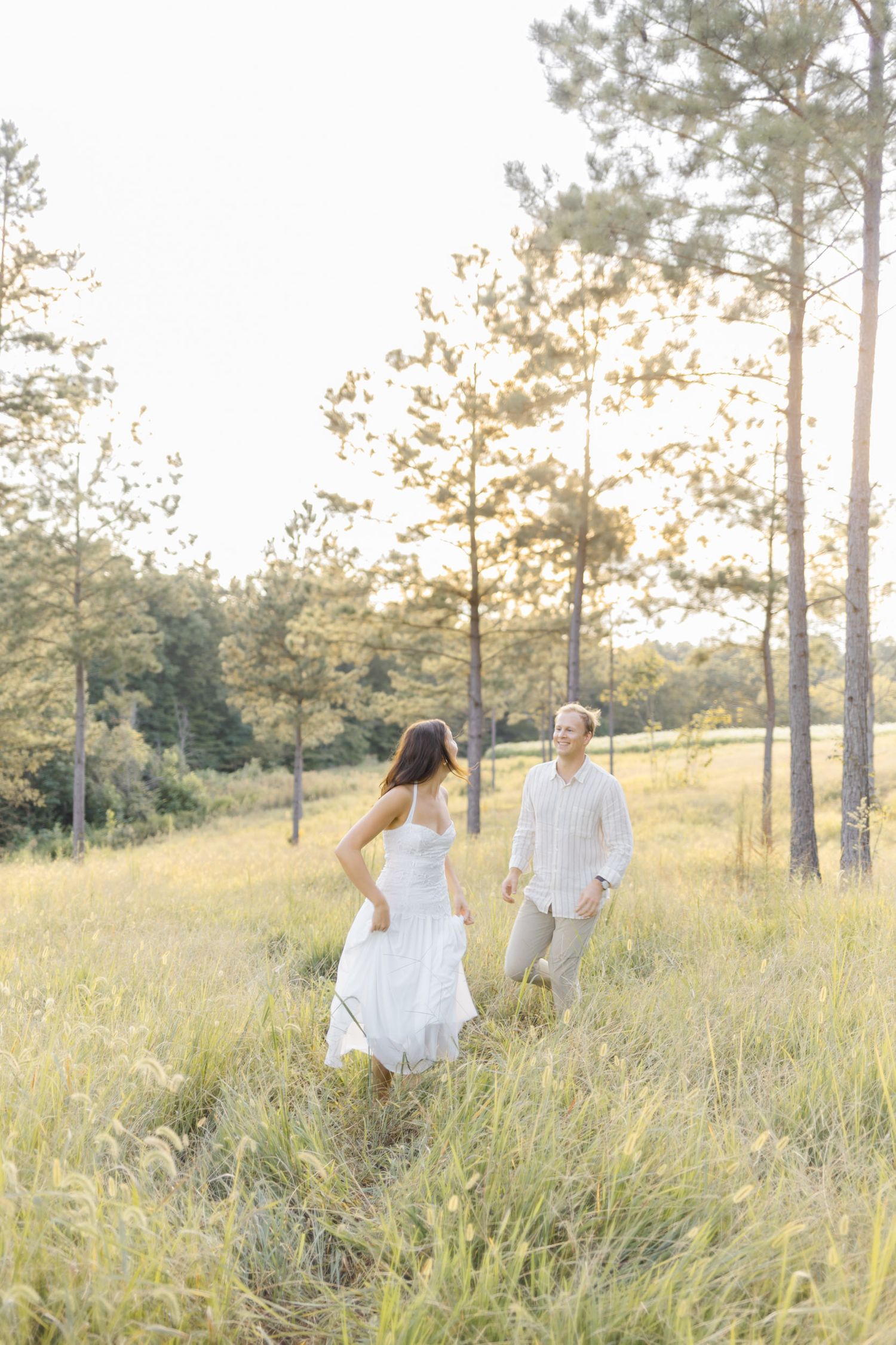 Couple dancing and twirling in a sun-drenched field with pine trees in the background during lifestyle portrait photoshoot.
