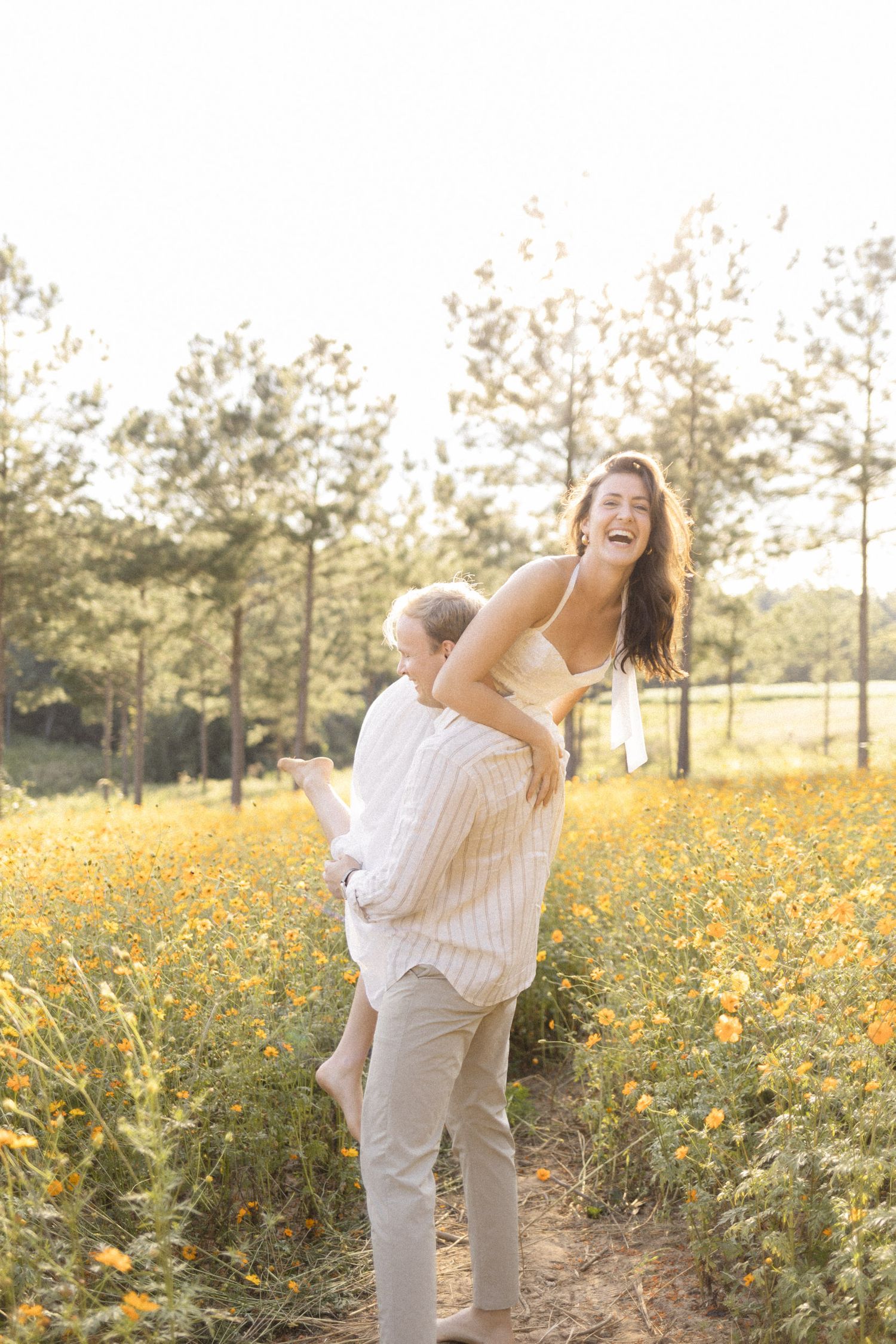 A joyful couple laughing in a sunny wildflower field, man lifting woman as golden blooms and pine trees surround them.