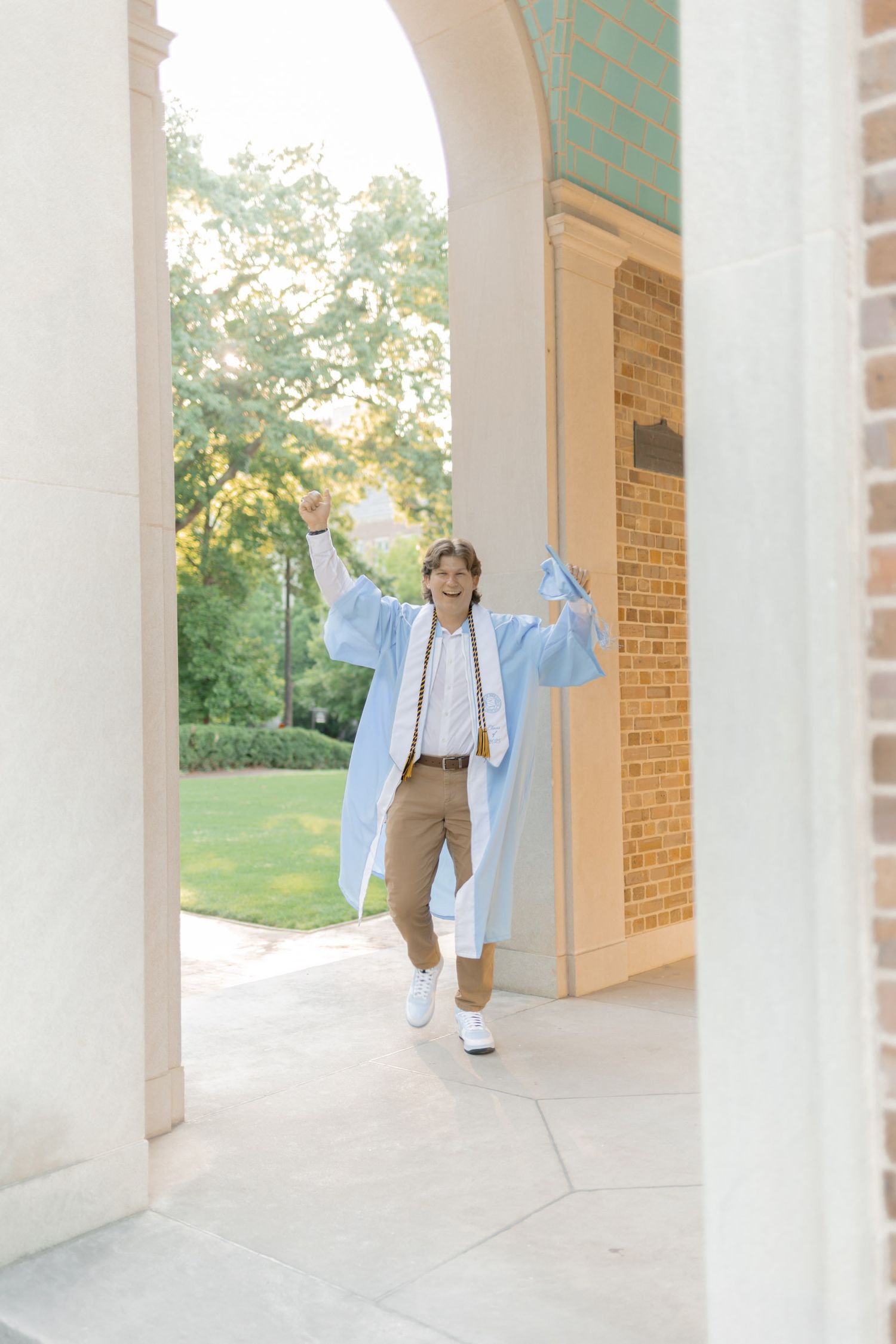 Excited graduate in light blue gown celebrating under campus archway with arms raised.
