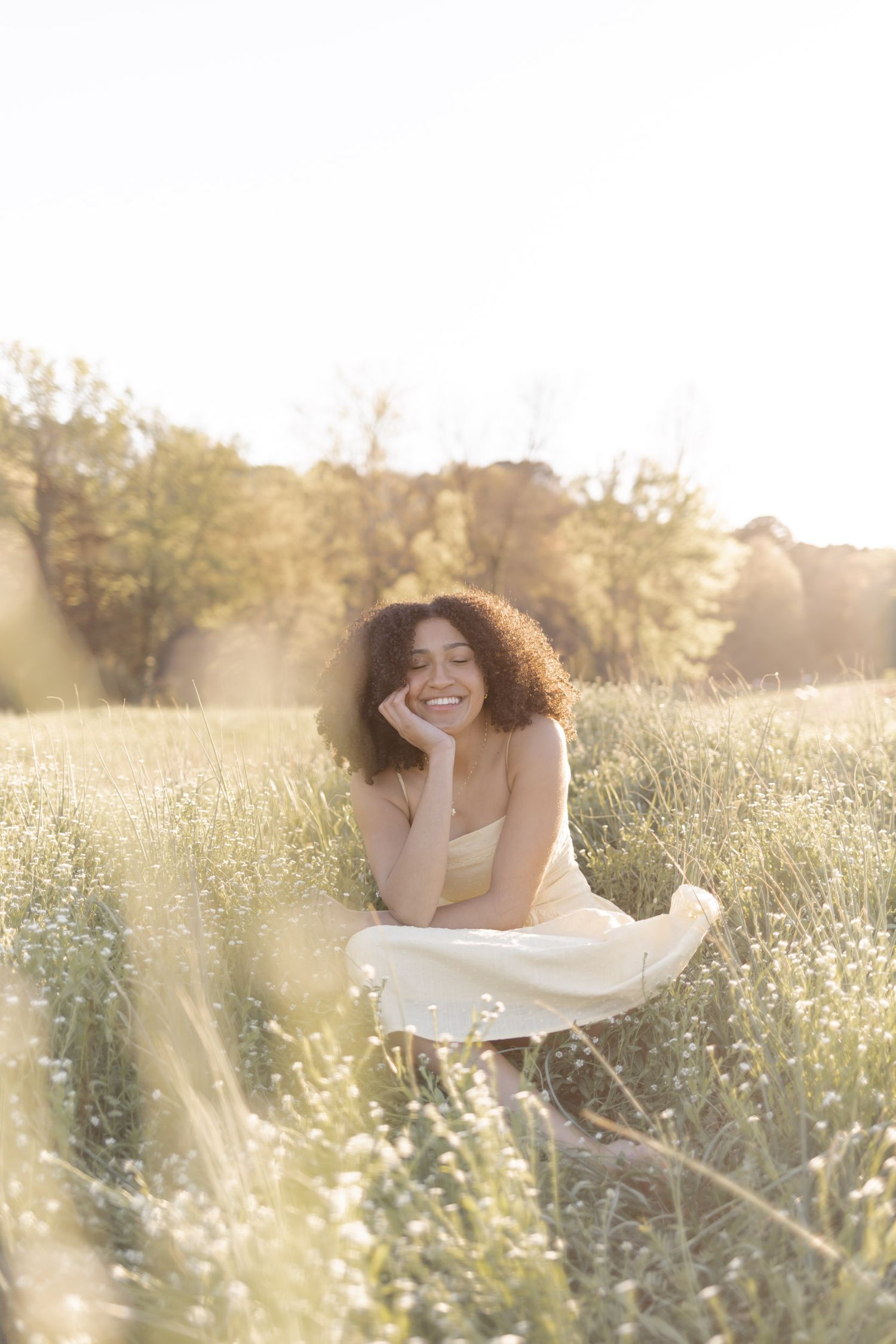 Young woman with curly hair smiling in a sunlit meadow wearing a white dress.