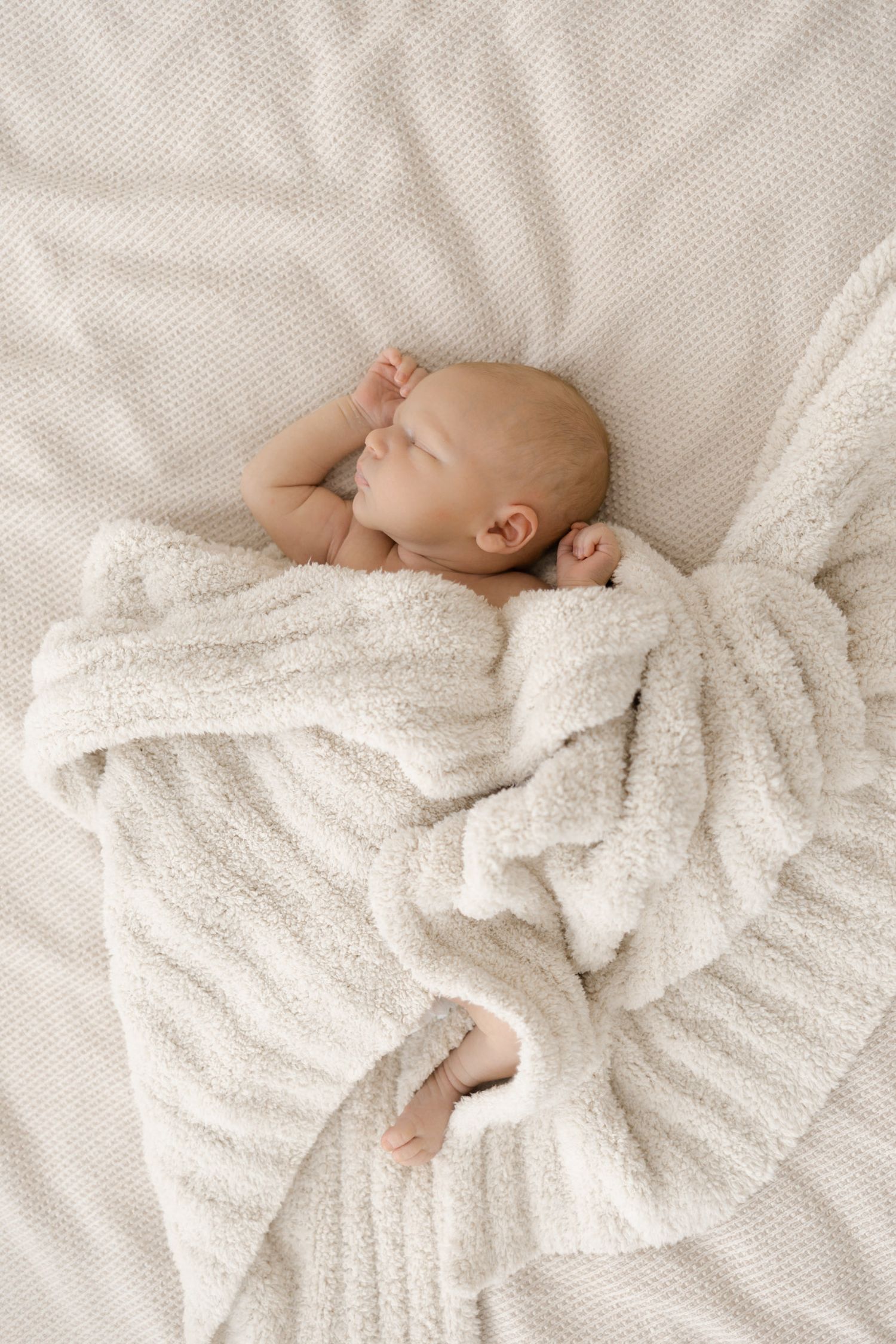 Peaceful newborn baby sleeping wrapped in soft white textured blanket on cream knit surface.