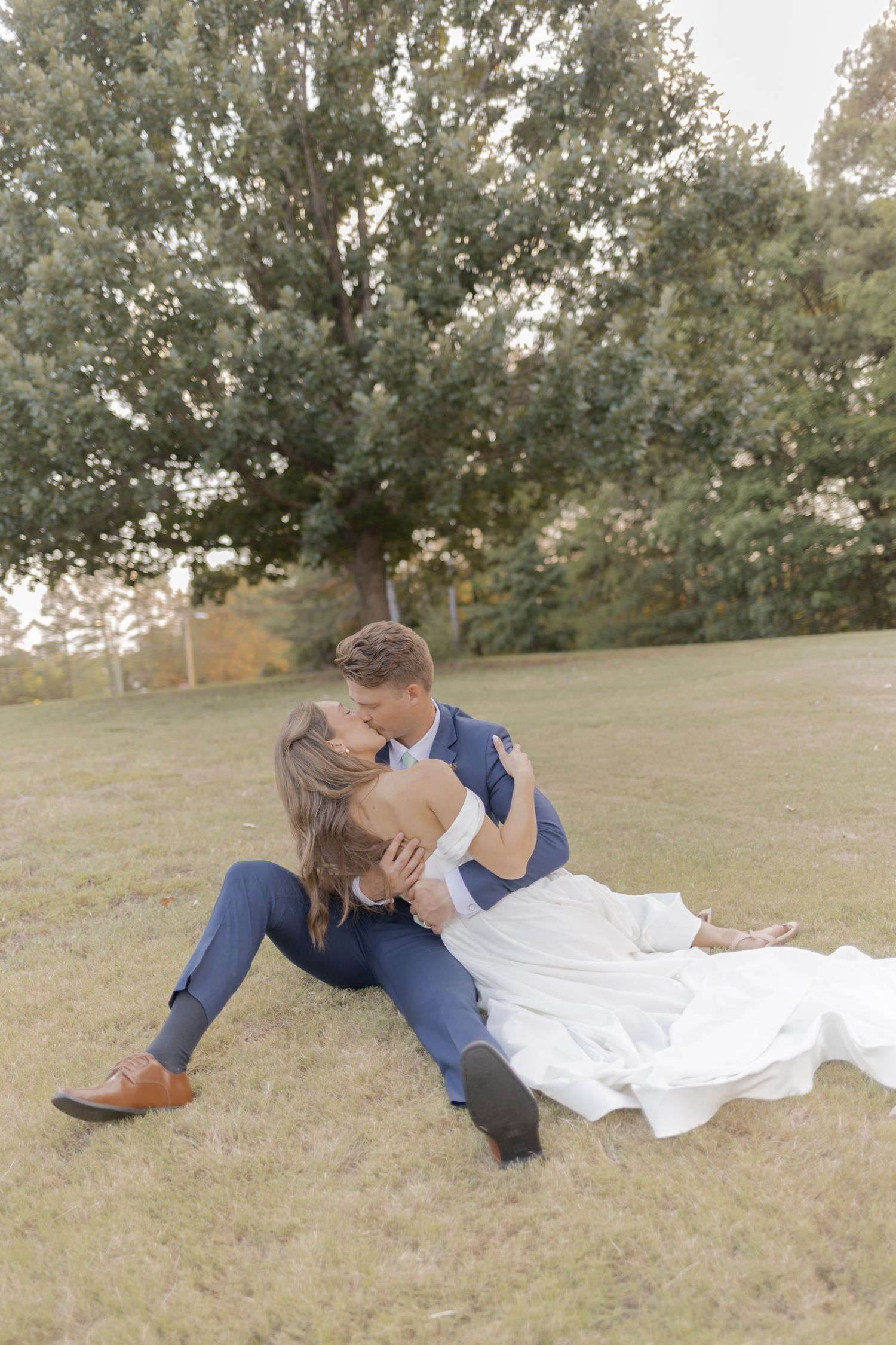 Bride and groom sharing a romantic kiss while sitting on green grass with trees in the background at outdoor wedding.