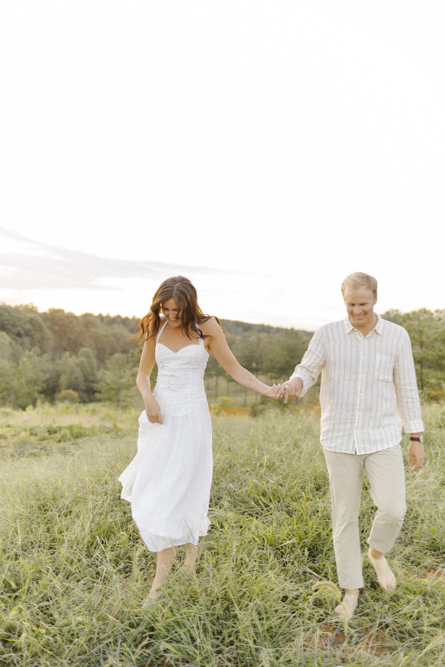 A couple holds hands while walking through tall grass in a summer field during engagement session in Chapel Hill, NC.